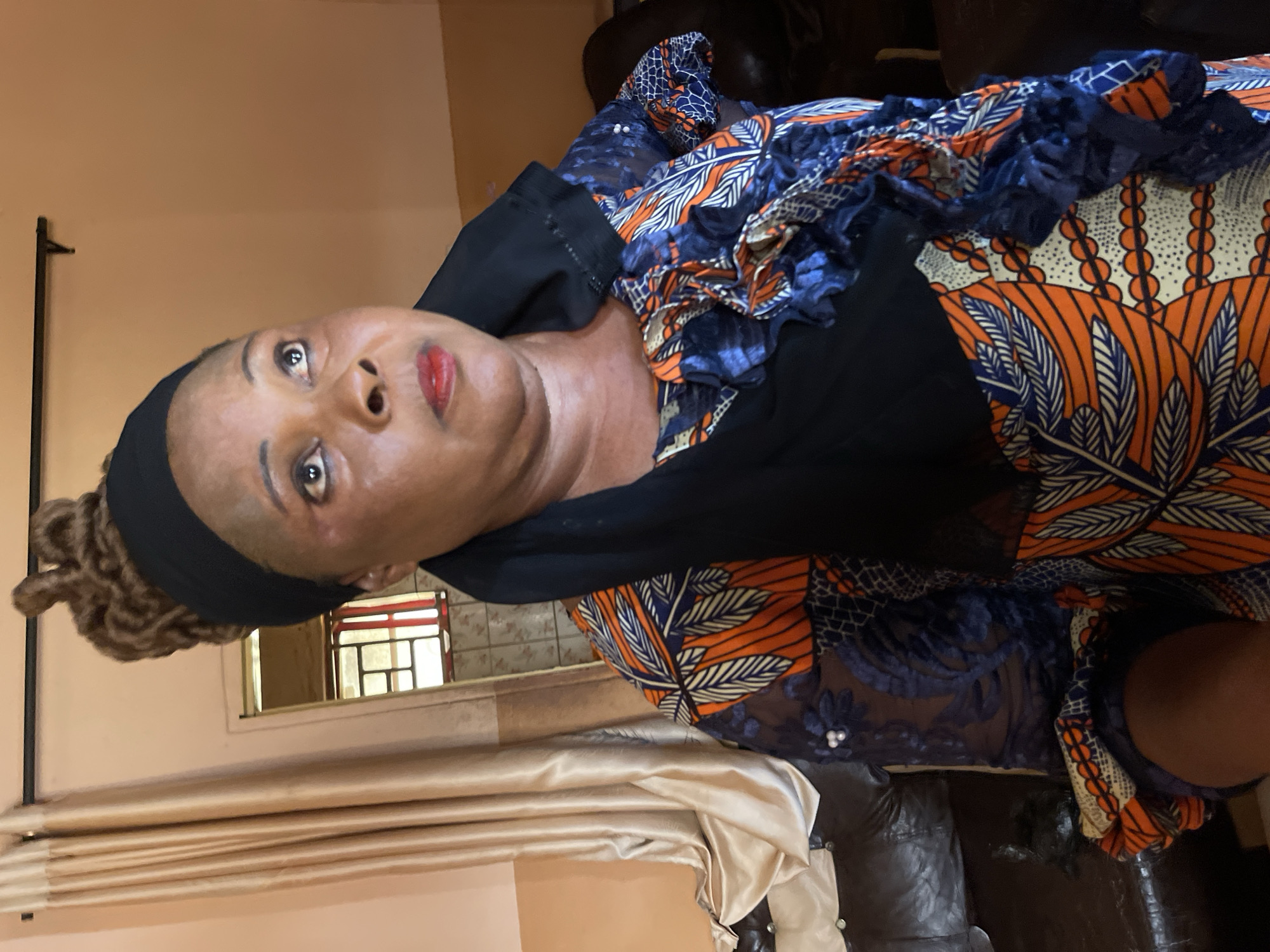 A woman is indoors, possibly in Muchiya, Nigeria. She has dark skin, brown eyes, and red lipstick. Her hair is in light brown dreadlocks, held back by a black headband. She is wearing a dress with an orange, white, and blue pattern with a navy blue ruffled top. The room has beige walls, curtains, and a window with a red and white pane design. A dark leather couch is visible in the background.