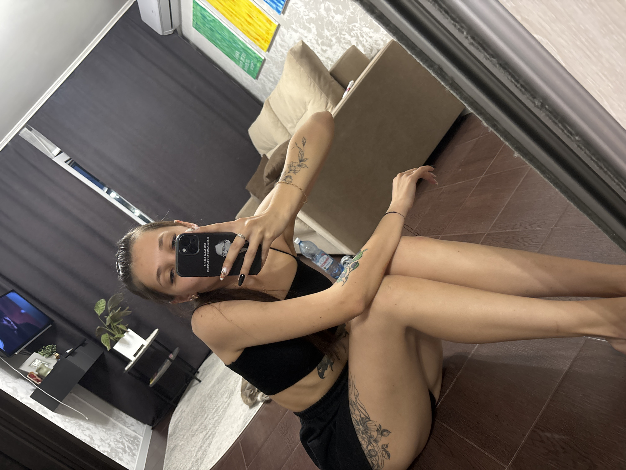 A woman with long brown hair is taking a selfie in a mirror. She is wearing a black crop top and black shorts. She has tattoos on her left thigh and left arm. She is holding a black iPhone in her right hand. In the background, there is a couch, a television, and a plant. The floor is made of dark brown tiles. The scene appears to be an indoor living space. The image is not geographically specific to Novokuznetsk, Russia.