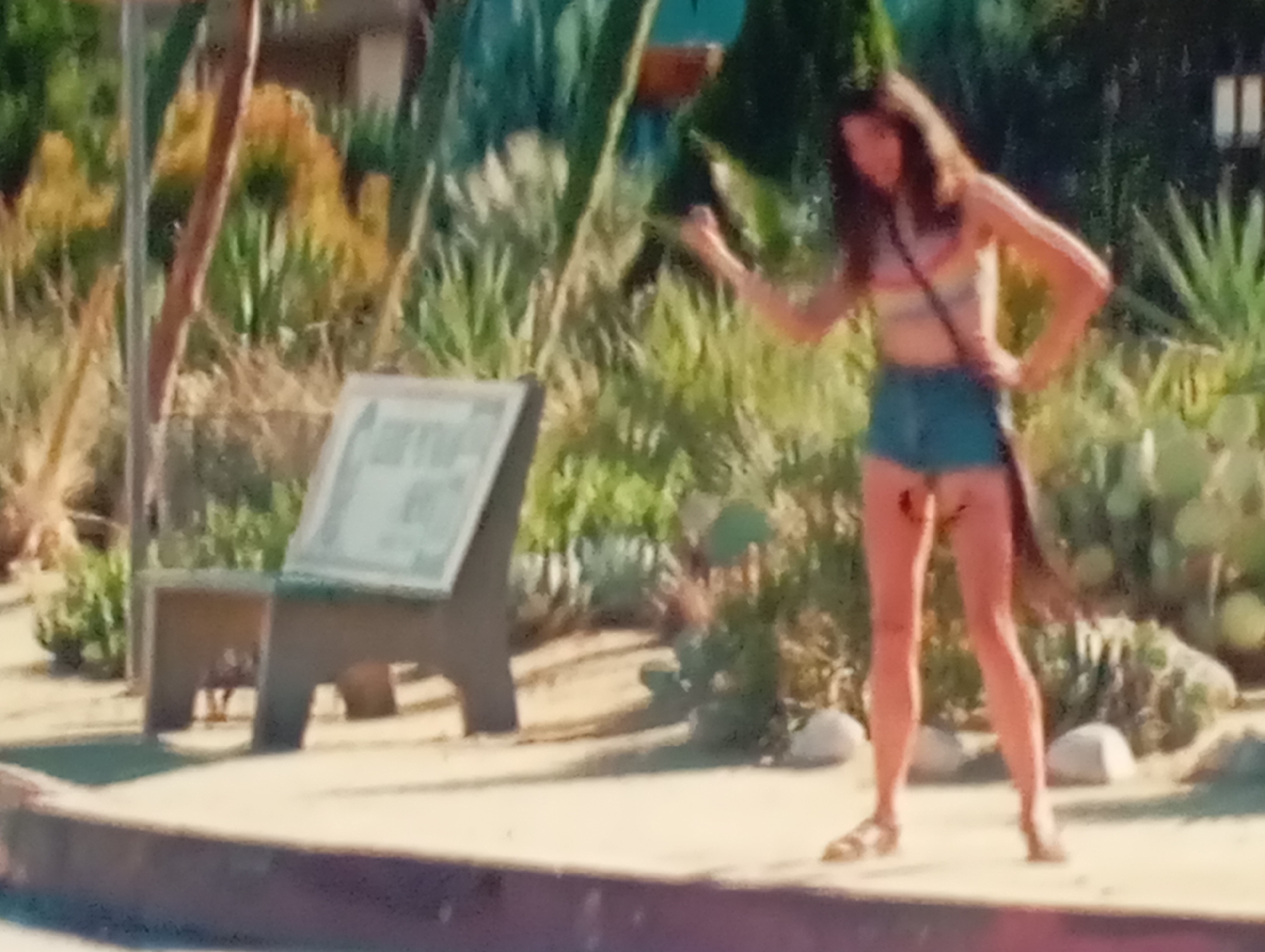 The image shows a young woman with long, dark hair, wearing a striped tank top and denim shorts. She is standing outdoors in what appears to be a sunny location, with lush green foliage and some arid-looking plants in the background. To her left, there is a dark wooden signpost with a white sign on it. The woman is posed with her hands on her hips, looking off to the side with a relaxed expression. The lighting suggests it is daytime, possibly afternoon, with the sun casting shadows. The overall scene is casual and summery, implying a relaxed outdoor setting. There is no visible text in the image that can be deciphered. The location context provided for Sofiivska Borshchahivka, Ukraine, suggests the image was taken in that region.