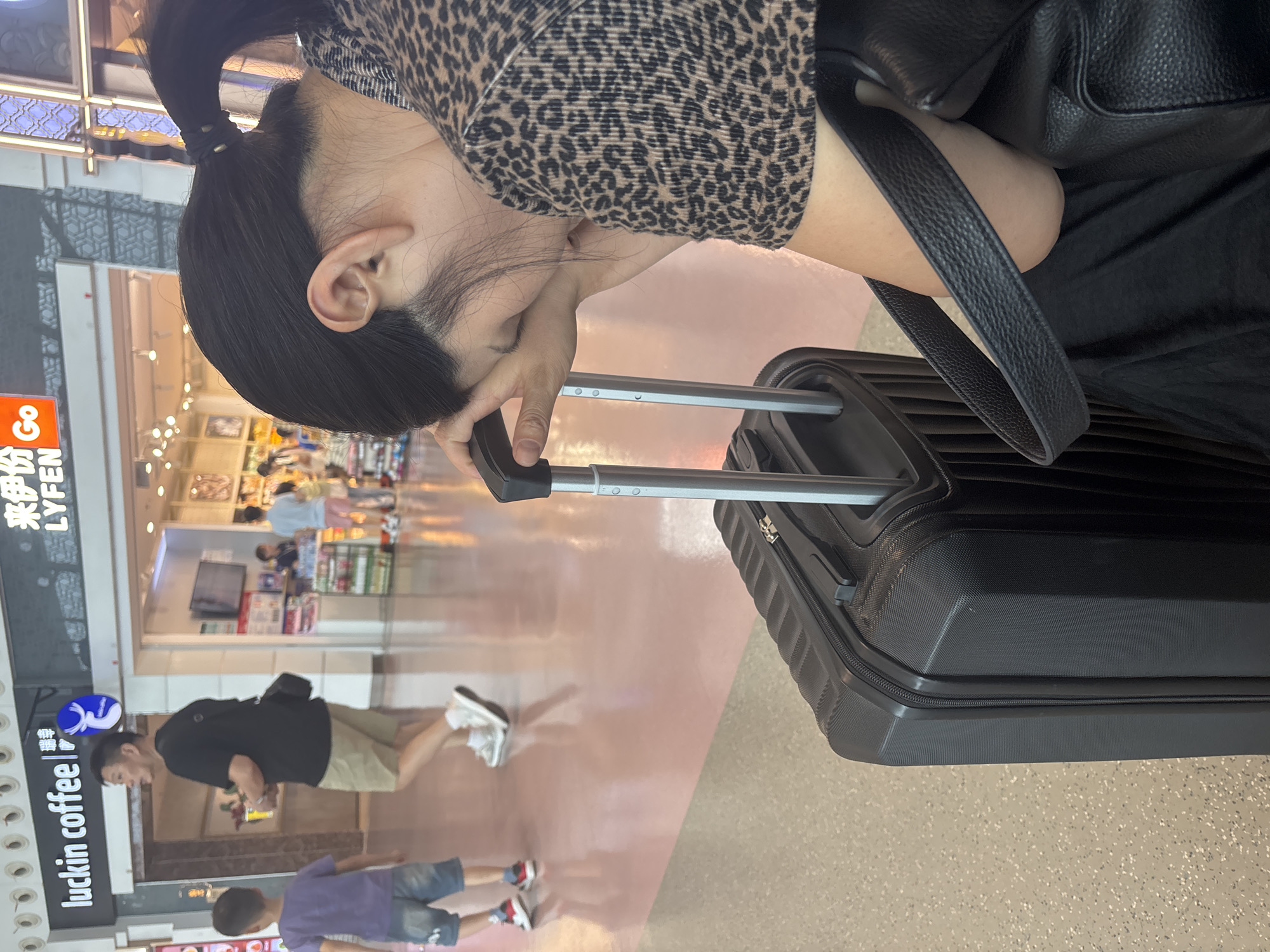 A woman with dark hair pulled back in a ponytail, wearing a leopard print top and a black skirt, sits with her head resting on her hands, which are placed on the handle of a black rolling suitcase. She appears to be distressed or tired. Beside her is another black rolling suitcase. In the background, a man in a black t-shirt and khaki shorts walks away from the camera, looking over his shoulder. A young boy in a purple t-shirt and denim shorts walks in front of the man. Behind them are shop fronts, including one labeled "luckin coffee" and another with Chinese characters and the word "Go". The floor is a pale pink tiled surface. The scene appears to be an indoor public space, possibly an airport or a shopping mall in Xinwu District, China.