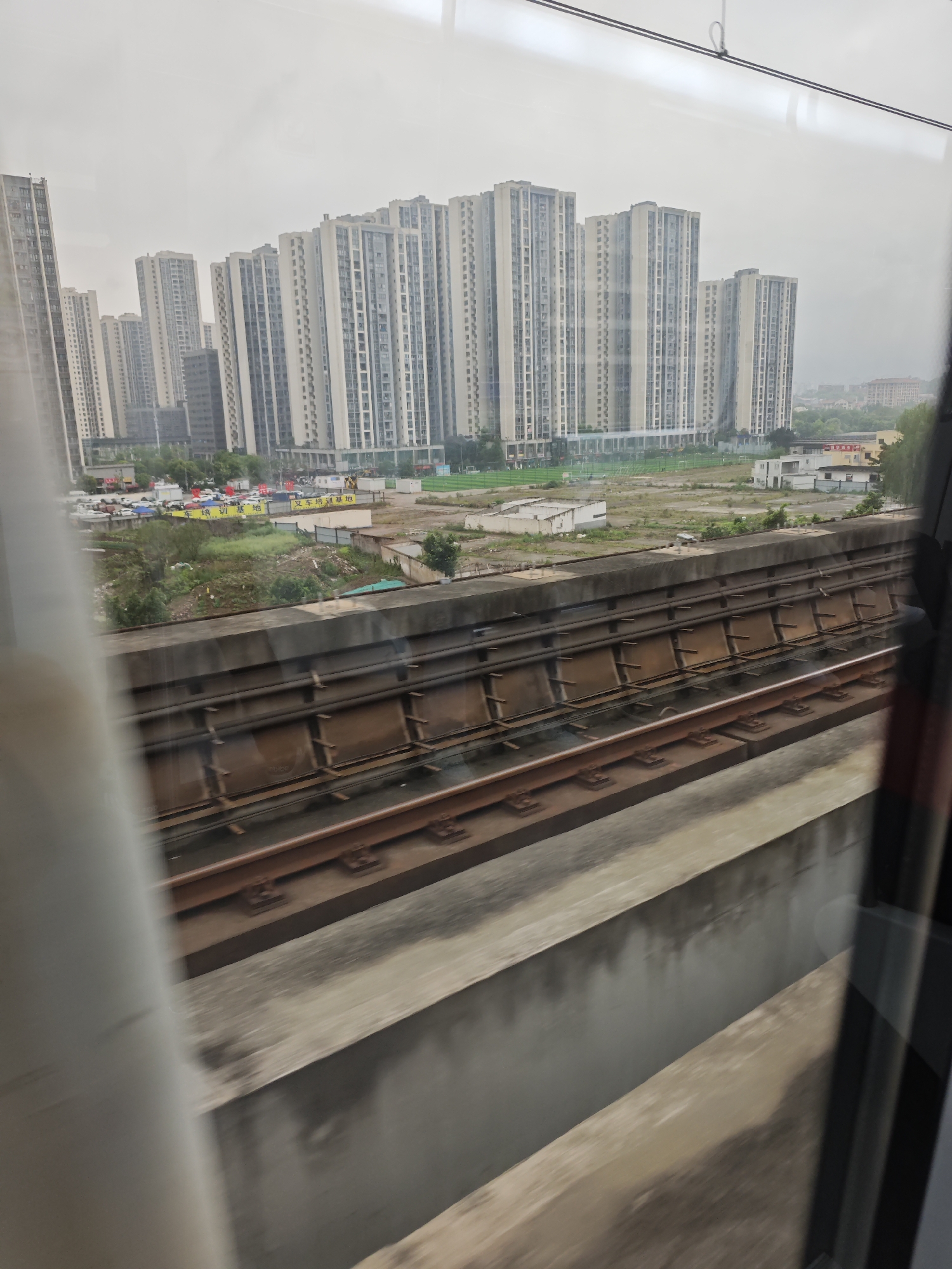 The image, taken from inside a moving train in the Chongqing High-tech Zone, China, shows a view through the window. In the background, a dense cluster of tall apartment buildings dominates the skyline under a cloudy sky. Below the buildings, there is a low-lying area with some greenery, scattered structures, and parked vehicles. The primary focus is on the elevated train tracks immediately outside the window, featuring the train rails and the supporting concrete structure. The window frame is partially visible on both sides of the frame, indicating the perspective from inside the train.