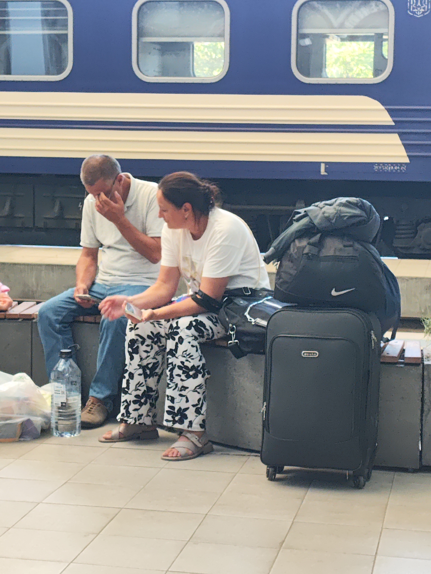 Two individuals, a man and a woman, are seated on a bench in front of a blue and white train. The man, on the left, is wearing a light-colored polo shirt and blue jeans, and he is touching his face. The woman, on the right, is wearing a white t-shirt and patterned pants, and she is looking at her phone. In front of them, a black suitcase and a black duffel bag are stacked, with a jacket draped over the duffel bag. A clear plastic bottle of water is on the ground to the left of the bench, along with a plastic bag. The ground is tiled. The scene appears to be a train station platform. The background shows the side of a train with windows. There is no specific indication of the city or country, however, if this image were to be associated with Chișinău, Moldova, it would likely be at the Chișinău railway station.