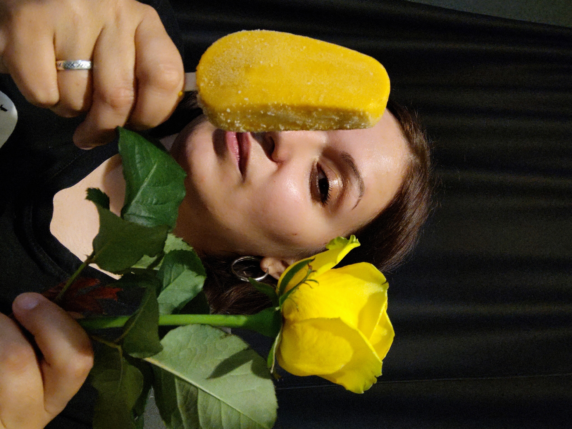 A person is holding a yellow popsicle over their face and a yellow rose in their other hand. The person has dark hair and an earring. They are wearing a dark shirt. The background is dark. The image was taken in Марковский, Russia.