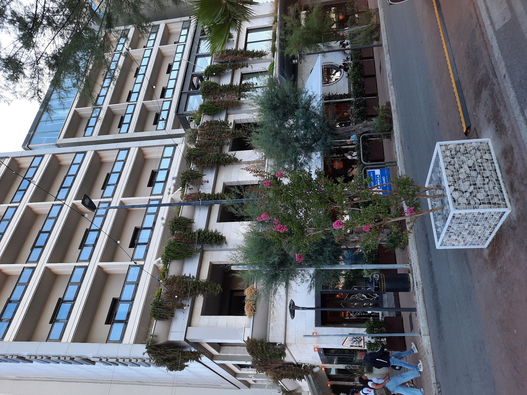 This image depicts a street scene in Pesaro, Italy, with a prominent modern hotel as the backdrop. The hotel is a multi-story building with a light-colored facade and numerous balconies, many of which are adorned with hanging plants, giving the structure a lush, green appearance. On the ground level of the hotel, there is a restaurant or cafe with outdoor seating, indicated by tables and chairs visible under a white canopy.
In the foreground, the street is paved with asphalt and a pedestrian walkway runs alongside. Several people are walking on the sidewalk, including a group of three individuals on the left side of the frame wearing casual clothing. A tall street lamp stands on the walkway. A white, cube-shaped planter containing white stones and a small tree with pink flowers is situated in the lower right portion of the image.
In the mid-ground, trees and foliage are present, with a larger tree with pink flowers positioned centrally and to the right, partially obscuring the hotel. A black trash receptacle is located near the entrance of the building on the left. Signs are visible on the building's facade, some with text. The overall atmosphere suggests a pleasant day in a coastal city, with the hotel and its greenery creating a striking visual against the urban environment.