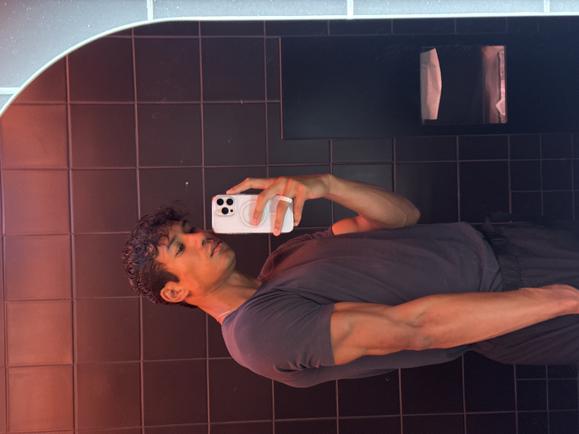 A young man is captured taking a mirror selfie in an indoor setting, likely a restroom or changing room. He is shown from the chest up, holding a white smartphone horizontally with both hands, positioned to photograph his reflection. He has dark, curly hair and is wearing a dark grey short-sleeved t-shirt. His muscular build is noticeable in his arms.
The background features dark, square-tiled walls. Above the man's reflection, a ceiling with light grey or white tiles is visible, featuring a distinct curved edge on the left side. On the right side of the image, behind the man's head, a toilet paper dispenser holding two rolls is mounted on the tiled wall; one roll appears partially used and folded.
The scene is lit with a prominent reddish-orange light source coming from the left, casting a warm glow on the man's face, neck, and right arm, creating a strong contrast with the dark background tiles. Overhead lighting is also present, reflected in the ceiling. The man maintains a neutral expression, looking directly at the phone. No visible text appears in the image.