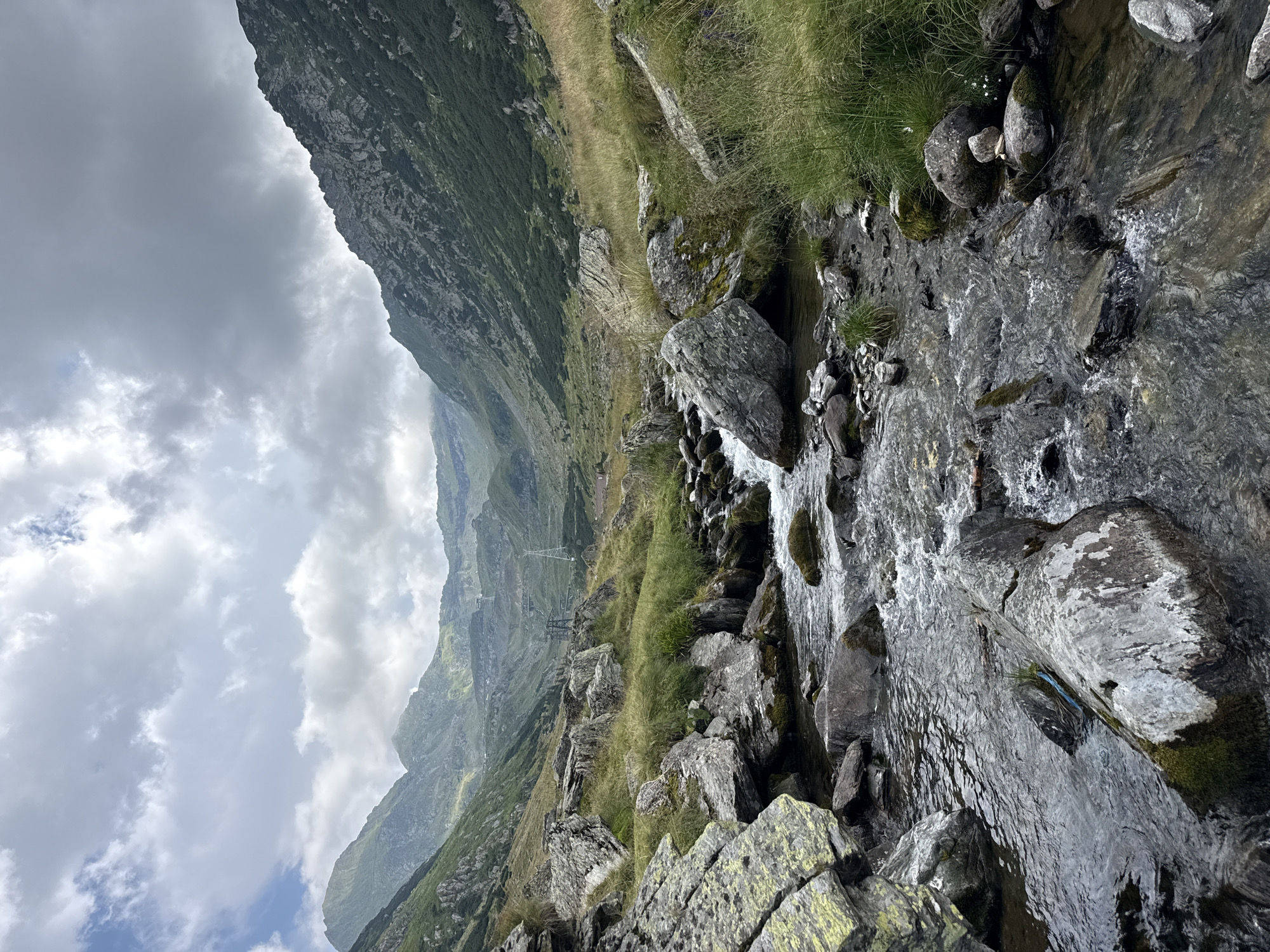 The image depicts a mountain landscape with a rocky stream flowing through it. The stream occupies the foreground, with its water cascading over various-sized rocks. The banks of the stream are lined with large boulders and overgrown with green grass and moss. In the mid-ground, the terrain slopes upward, revealing more rocks and vegetation, and further in the distance, a cable car system is visible ascending a steep mountain slope. The background consists of a majestic mountain range, with peaks partially obscured by clouds. The sky above is a mix of gray clouds and patches of blue, suggesting an overcast or partially cloudy day. The overall scene is a natural, rugged environment, likely within the Transfăgărășan highway area near Cârțișoara, Romania, known for its dramatic mountain scenery and infrastructure like cable cars. No people are visible in the image.