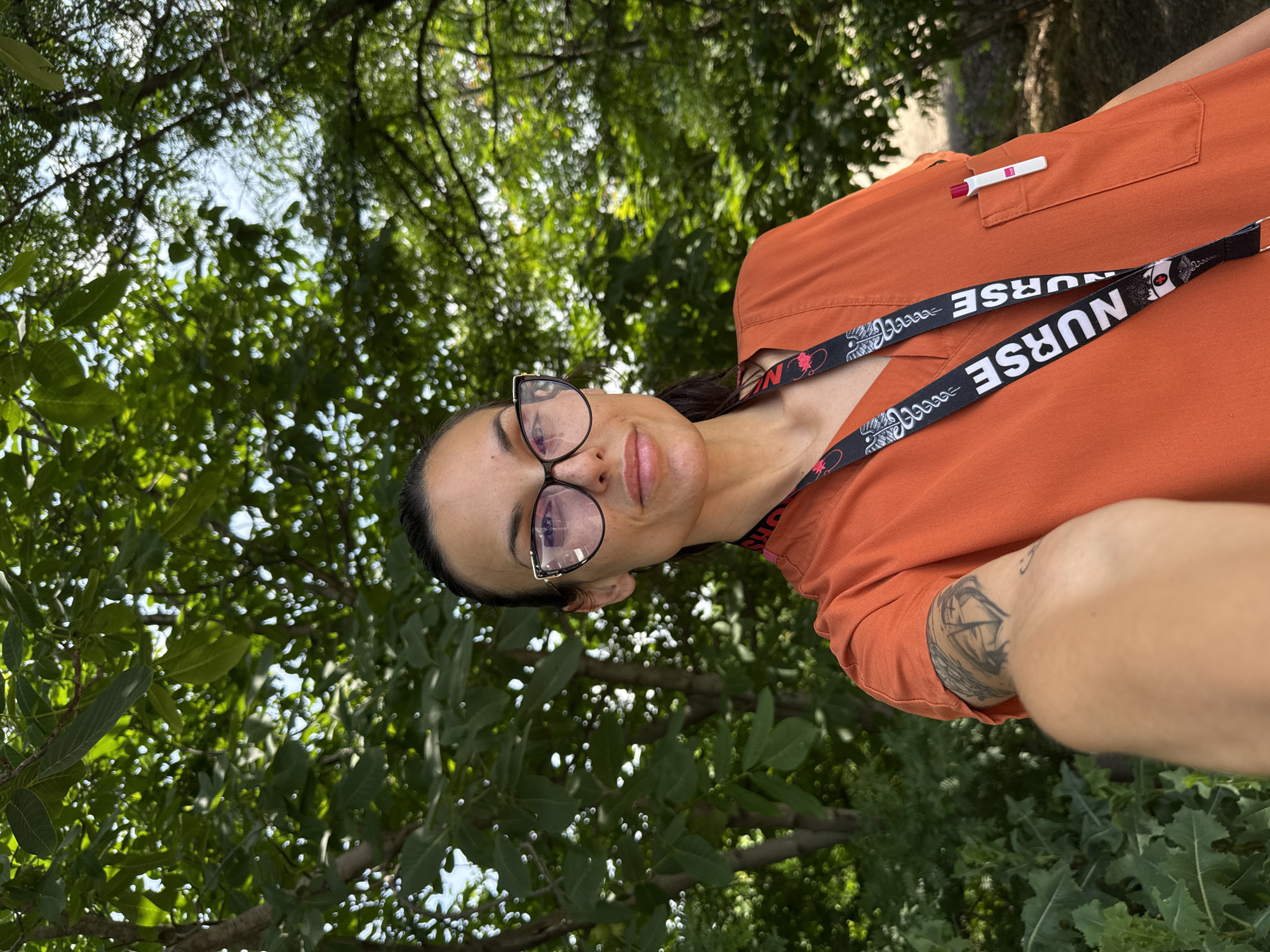 An adult female is depicted in a selfie-style portrait taken outdoors, presumably within the city of Pleven, Bulgaria. She has dark hair pulled back and wears black and gold-framed eyeglasses. She is dressed in an orange V-neck scrub top. Around her neck is a black lanyard featuring multiple instances of the word "NURSE" in white text, interspersed with red and white caduceus symbols. A white pen with a red top is clipped into the left chest pocket of her scrub top. A tattoo with geometric or abstract designs, including what resembles a heart shape, is visible on her left upper arm. The background consists of dense green foliage from trees and bushes, with glimpses of blue sky visible through the leaves, indicating a bright, sunlit environment.