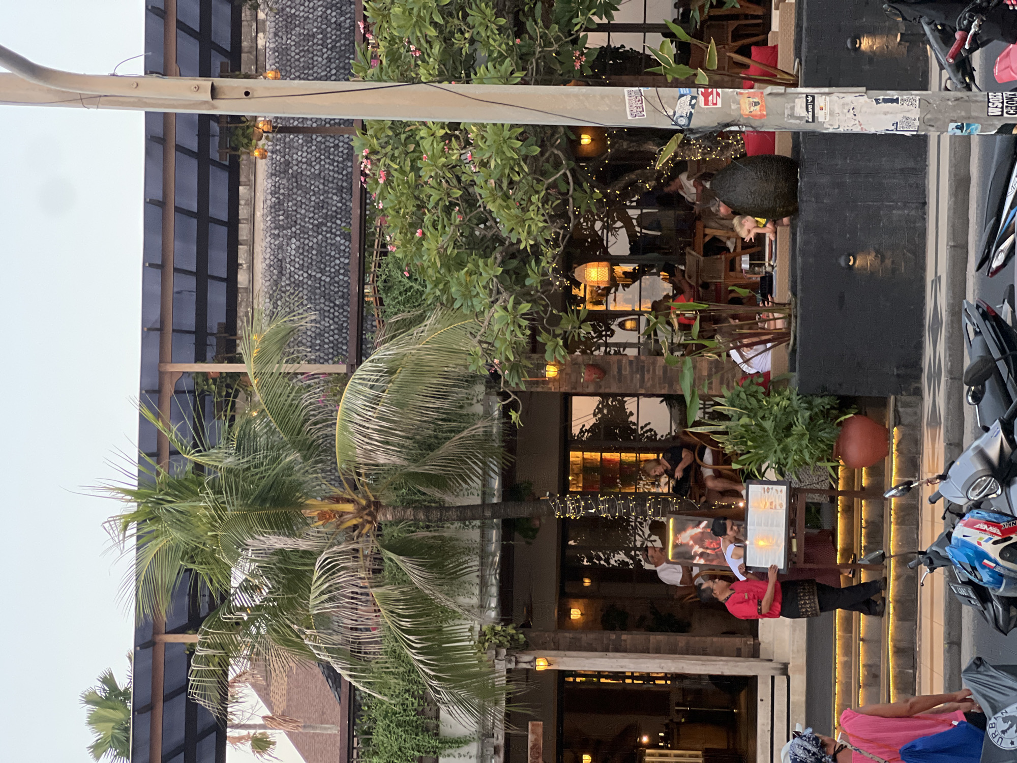 The image depicts a street-level view of a restaurant or cafe in Kuta, Indonesia, at dusk. A prominent palm tree with lush fronds stands in the foreground, partially obscuring the entrance to the establishment. The building features a multi-level structure with a covered outdoor seating area illuminated by string lights and ambient lighting from within. Several patrons are seated at tables, some engaged in conversation and others perusing menus. A server, dressed in a red shirt and black pants with a patterned waist sash, stands by a display stand presenting a menu to a seated couple. A woman in a pink top and headscarf is walking away from the camera down a set of illuminated stairs. In the foreground, several motorcycles are parked along the sidewalk. The overall atmosphere suggests a lively evening scene in a popular tourist destination.