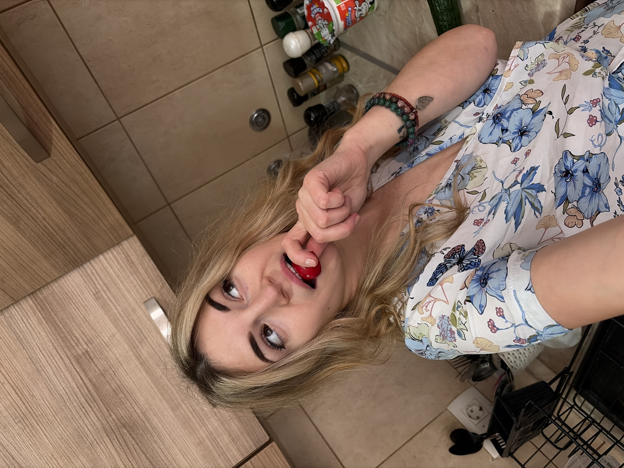 The image displays a kitchen interior located in Budva, Montenegro. A female subject, with blonde, wavy hair, is positioned centrally. She wears a white dress adorned with blue, orange, and green floral and butterfly patterns. Her right hand holds a small red, round object, consistent with a cherry tomato, to her mouth. Her eyes are directed upwards and to the right. On her left wrist are multiple beaded bracelets, one featuring a heart-shaped charm, and a small tattoo is visible on her forearm. The kitchen environment includes light wood-grain upper cabinets with a metal handle, a light brown tiled backsplash, and a light brown countertop. On the countertop's right side, several spice jars and a small white cup with red and green branding are visible. To the left, a black metal dish drying rack, a white utensil holder with various utensils, a black spatula, and a white electrical outlet are present.