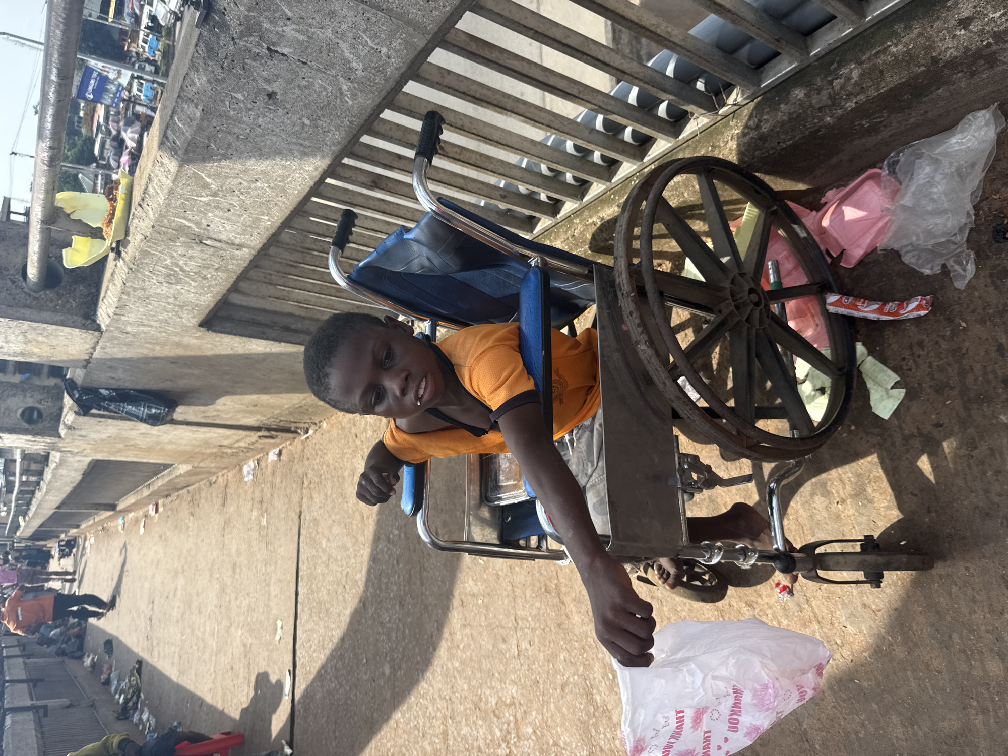 A young boy in a wheelchair is positioned on a bridge or elevated platform in Onitsha, Nigeria. He wears an orange shirt and gray pants and is holding a white plastic bag. The wheelchair is next to a metal railing. In the background, the scene is crowded with many people and various objects scattered along the walkway.