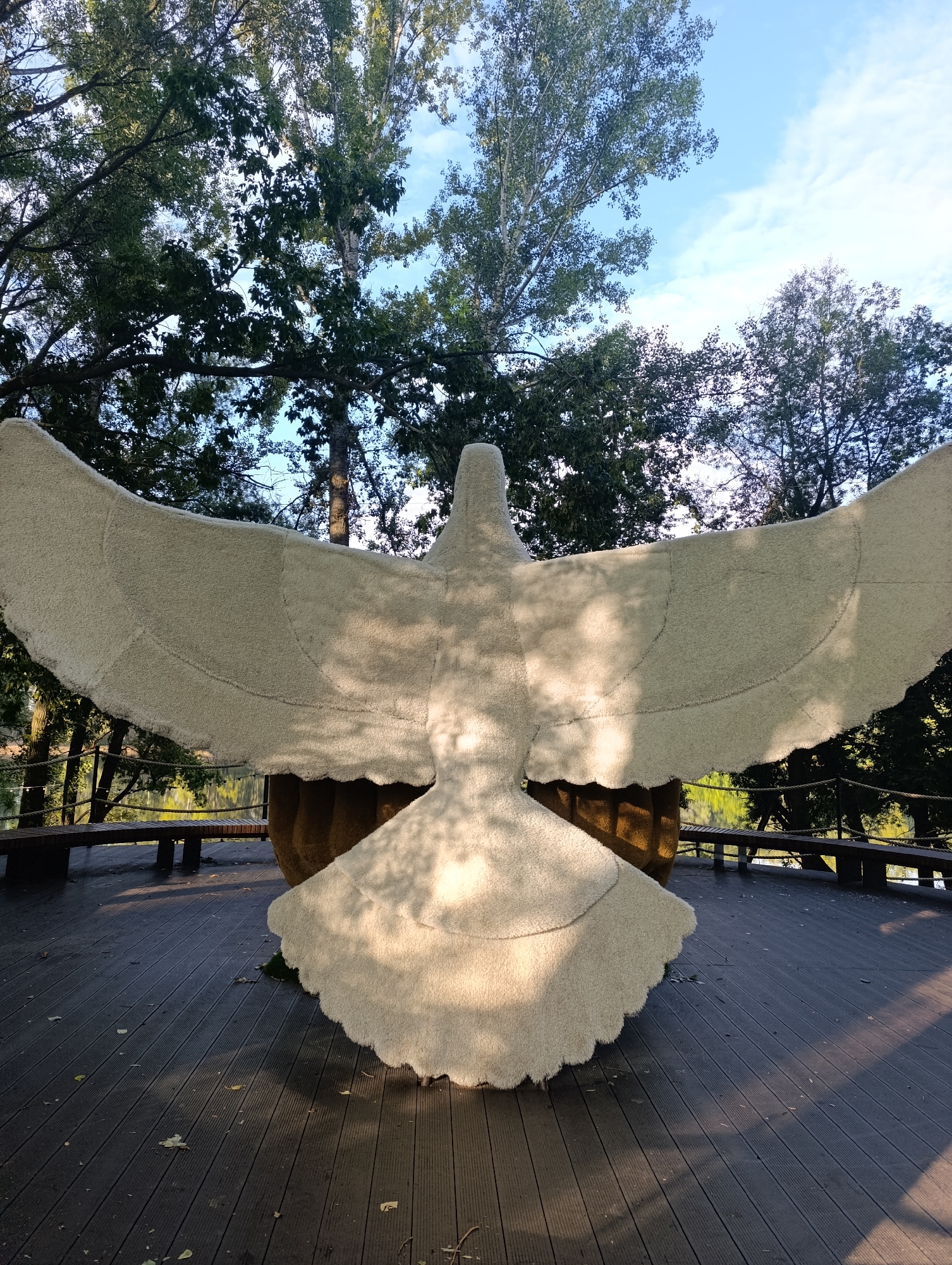 An outdoor daytime shot shows a large, fluffy, white sculpture of a bird with its wings spread wide. The sculpture is made of a fuzzy material and is situated on a dark wooden deck. Behind the sculpture, trees with green leaves and a blue sky with white clouds are visible. To the left and right of the sculpture, there are wooden benches and a railing. The sculpture's base is a large, brown, textured object. Sunlight casts shadows on the deck and the sculpture.