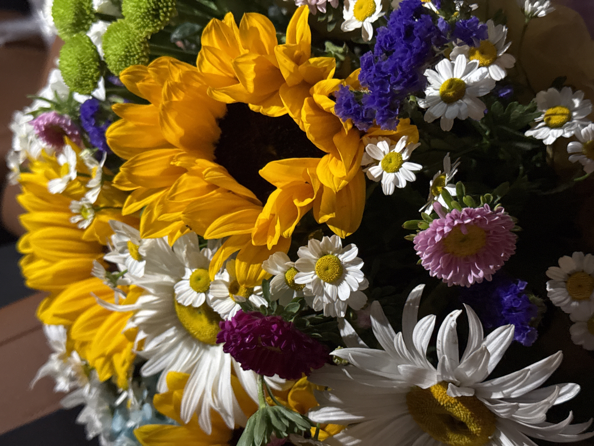 This media file is a close-up, brightly lit photograph of a vibrant bouquet of mixed flowers. The dominant subjects are sunflowers with their large yellow petals and dark centers, interspersed with white daisies featuring yellow centers. There are also clusters of small, bright purple flowers, and some soft green, spherical flowers. A few pink aster-like flowers add to the variety. The overall impression is one of a cheerful and abundant arrangement.
The setting appears to be indoors, with a dark, out-of-focus background suggesting the flowers are on a table or surface, possibly as a decorative centerpiece. There are no people or explicit location cues to indicate time of day, weather, or specific indoor location beyond the general domestic or decorative context. The lighting is direct and highlights the textures and colors of the petals and centers of the flowers. The arrangement is lush and full, with many flowers layered and overlapping. The colors are rich and saturated, with the yellow of the sunflowers, the white and yellow of the daisies, and the bright purple creating a striking contrast against the darker background. The focus is sharp on the main floral elements, drawing the viewer's attention to their intricate details. There is no visible text in the image.