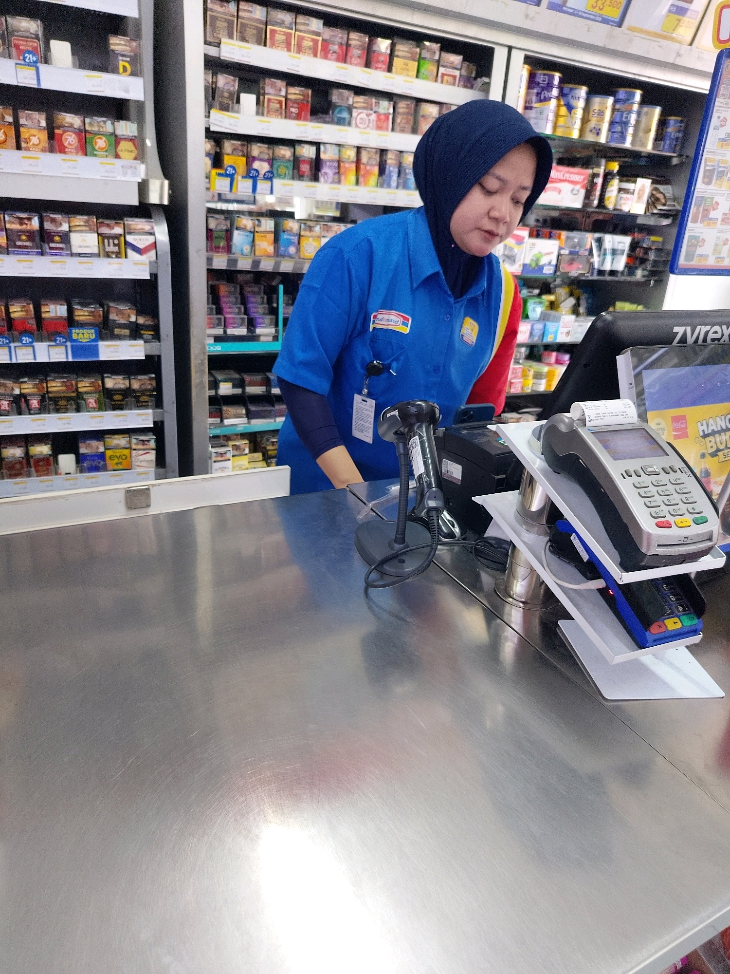 A woman wearing a blue uniform with a navy hijab and a yellow and red accent on her sleeve is standing behind a metal counter. She appears to be working at a convenience store, possibly a minimarket, in Mojokerto, Indonesia. The shelves behind her are stocked with various products, including cigarettes, canned goods, and other packaged items. In front of her on the counter are a barcode scanner, a receipt printer, and a credit card machine. The woman is looking down, possibly at the credit card machine or the products being scanned. The overall scene suggests a retail transaction in progress.