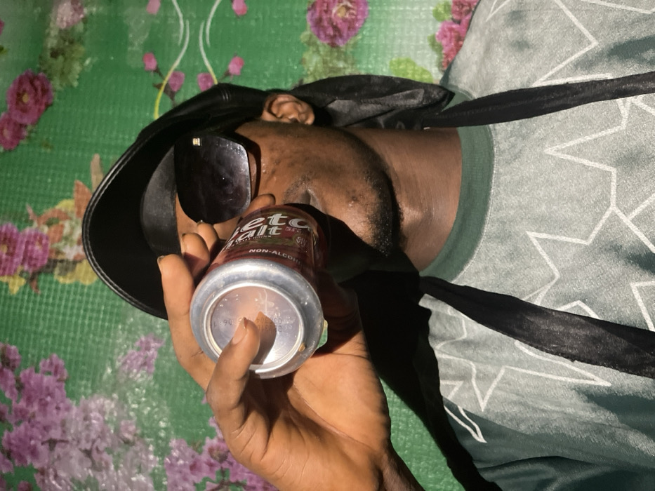 A person with dark skin, wearing sunglasses and a black head covering, is lying down. They're holding a can of "Beta Malt" non-alcoholic drink. The can is partially empty, showing the open top. The person's skin is slightly sweaty or wet. The background is a mix of green and floral patterns, possibly a tablecloth or blanket. A t-shirt with a white star pattern can be seen. The overall image is taken from a low, slightly angled perspective.