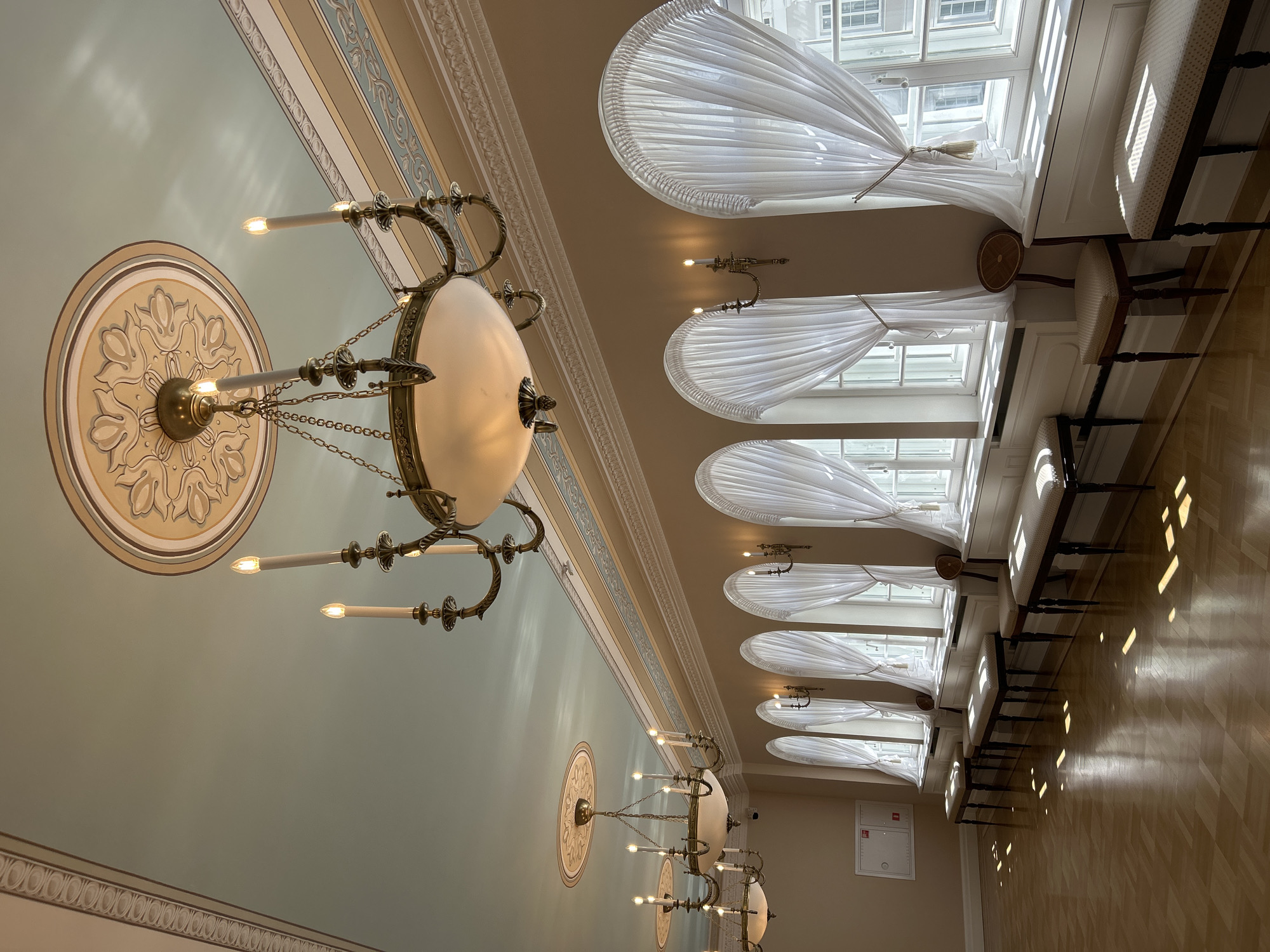 This image depicts an interior scene within a grand hall, likely a historical or government building, in the village of Dobasnenetski Selski Savek, Belarus. The room is spacious, with high ceilings adorned with decorative molding and ornate circular medallions. Several large, multi-tiered chandeliers with candle-like light fixtures hang from the ceiling, casting a warm glow. The walls are painted a neutral, warm tone, and are bordered by elaborate decorative trim.
Along one wall, a series of large, arched windows are covered with sheer white curtains that are gathered at the top and bottom. Sunlight streams through the windows, creating distinct patterns of light and shadow on the polished wooden floor. In front of the windows, a row of dark-legged benches with padded seats are neatly arranged. A single, ornate chair with a circular wooden back is placed between two of the windows.
In the background, a fire alarm and extinguisher cabinet is mounted on the wall. The overall atmosphere is one of elegance and historical significance, suggesting a well-preserved or restored interior space. There are no people visible in the image.