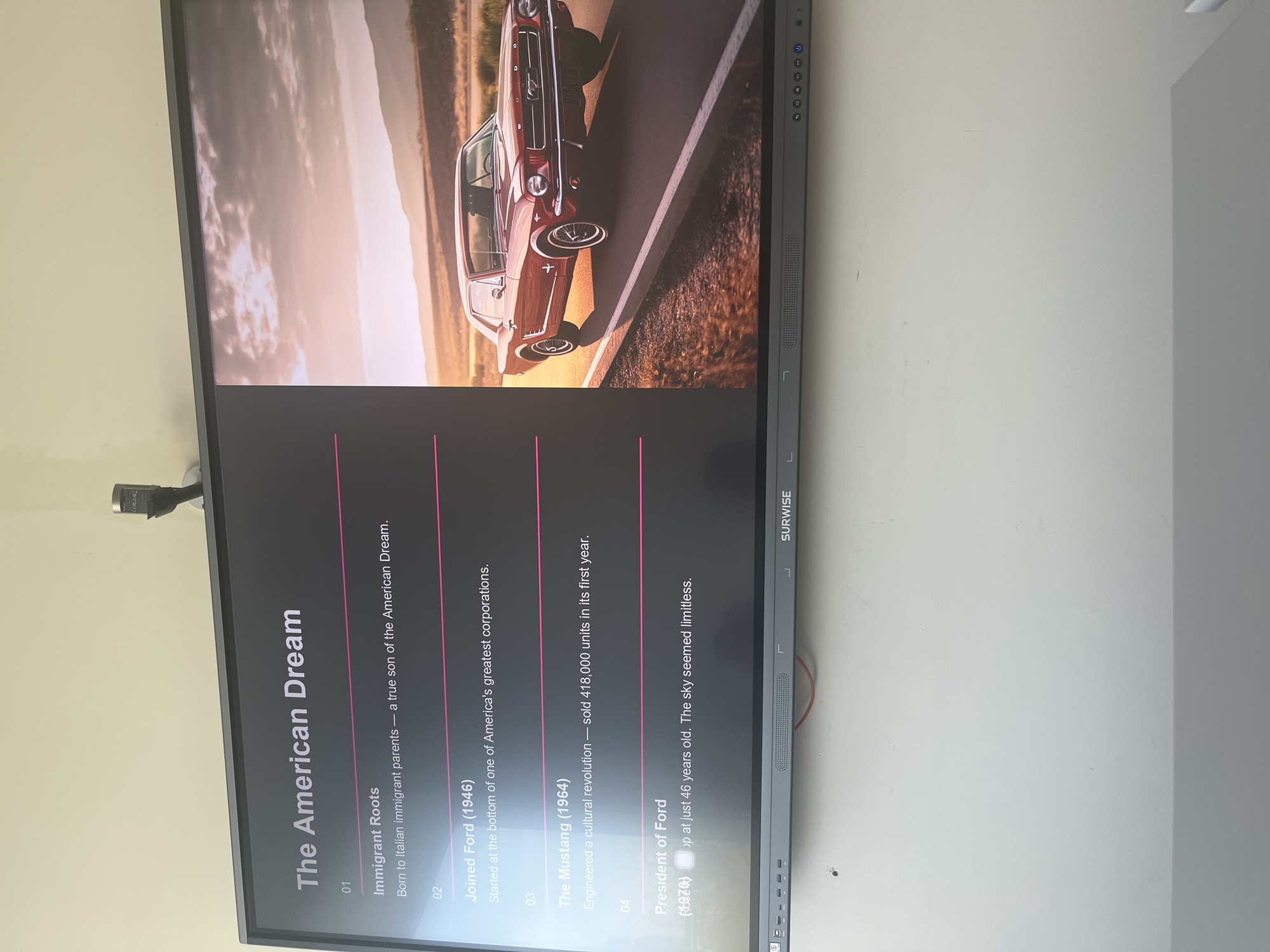 The image shows a flat screen TV mounted on a wall, displaying a presentation slide titled "The American Dream". The slide features a photo of a red Ford Mustang at the top. Beneath the image, there is text outlining a brief history of Ford, starting with "Immigrant Roots", continuing to "Joined Ford (1946)", "The Mustang (1964)" and ending with "President of Ford". The manufacturer's name "SURWISE" is visible at the side of the screen. The setting appears to be indoors, but the specific location is not identifiable, let alone Moscow, Russia.