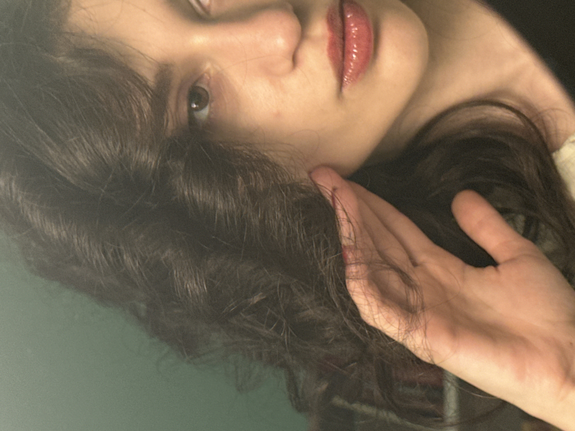 The image is a close-up, indoor photograph of a young person's face, with their hand gently touching their cheek. The person has dark, wavy hair that frames the right side of their face, partially obscuring their eye. Their lips are painted with a glossy, reddish-pink lipstick. The background is a plain, greenish-blue wall. There is no indication of the city of Sofia, Bulgaria, or any other specific location. The scene is static and lacks any narrative elements beyond the posed gesture of touching the face.