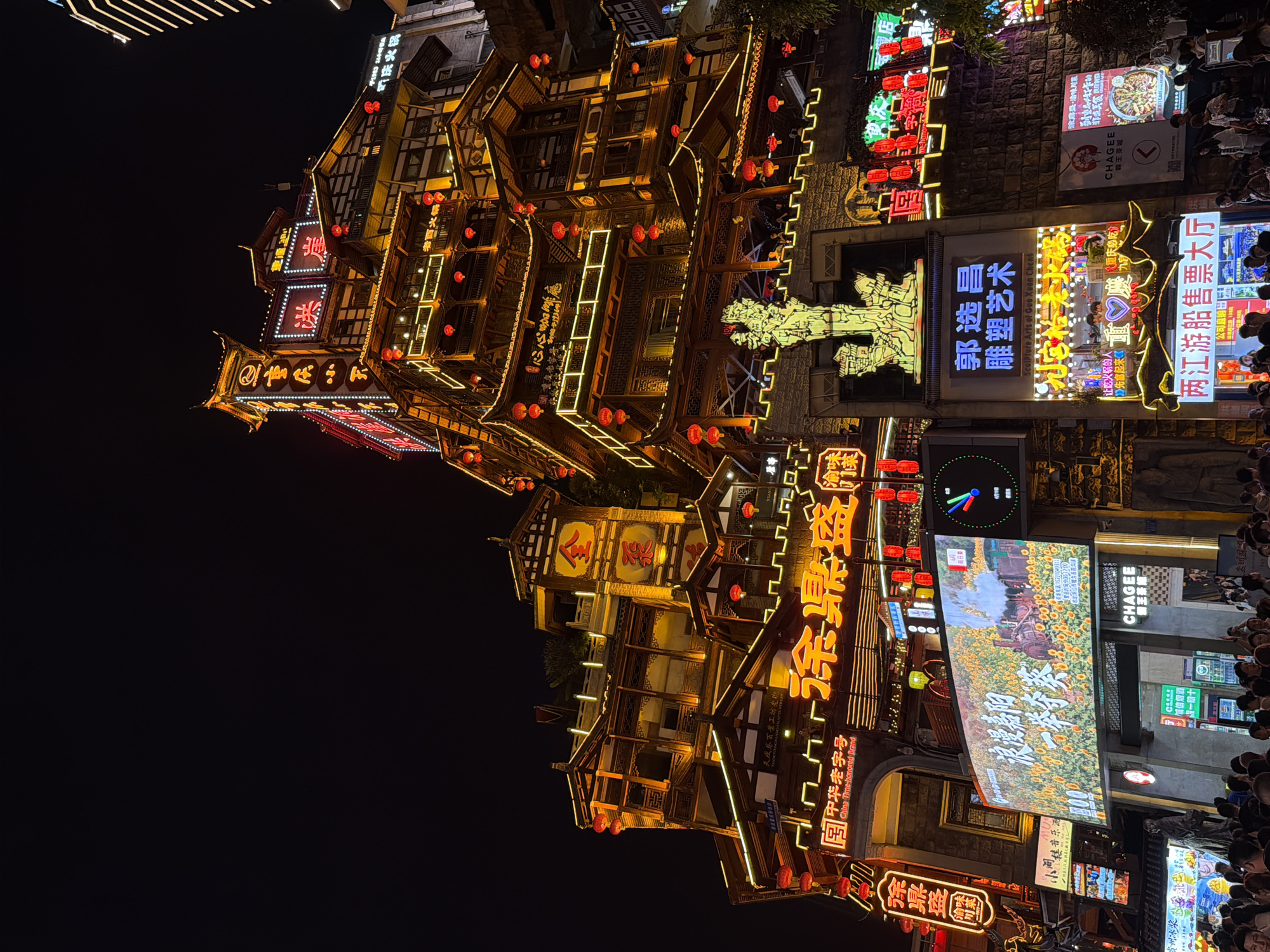 The image depicts a vibrant night scene in Yuzhong District, China, featuring a towering, multi-story building with traditional Chinese architecture illuminated by numerous golden lights and red lanterns. The building's facade is adorned with illuminated signage in Chinese characters, some of which are highlighted in neon. In the foreground, a large digital billboard displays a colorful image of a steam train amidst a field of sunflowers, with Chinese text above. To the right of the billboard, another illuminated sign reads "CHAGEE" with Chinese characters below. Below the billboard, a sign for "两江游船售票大厅" (Two Rivers Cruise Ticket Hall) is visible. A crowd of people is gathered at the base of the building, looking towards the illuminated structures and signs. The overall atmosphere is one of bustling urban nightlife and cultural display.