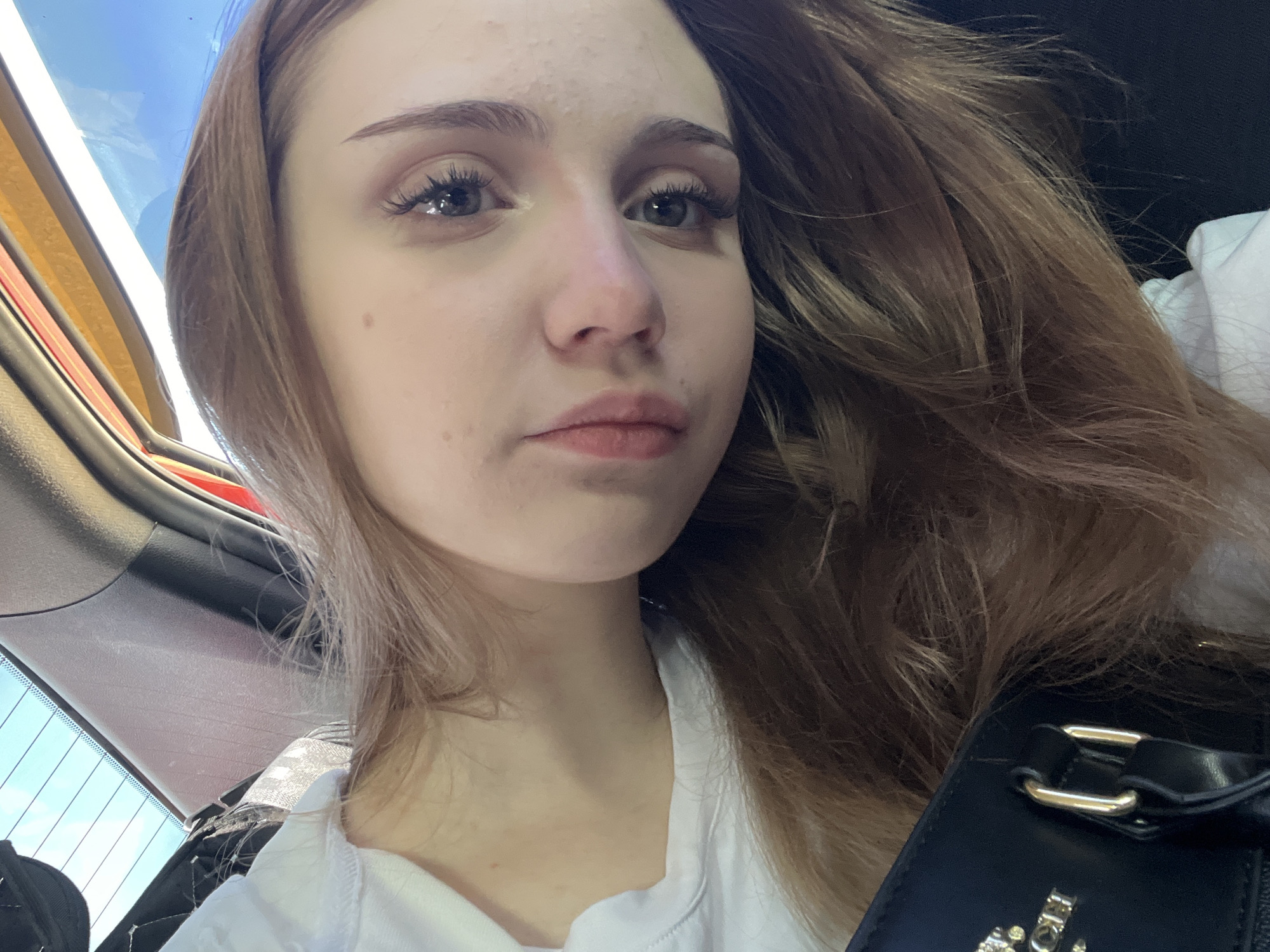 A close-up, low-angle shot shows a young woman with light brown hair tilted her head, looking towards the camera. She is wearing a white t-shirt and a black purse with gold hardware is visible in the foreground. The background appears to be the interior of a car, with sunlight filtering through the rear window. The image does not contain any information about the city of Марьевка, Russia.