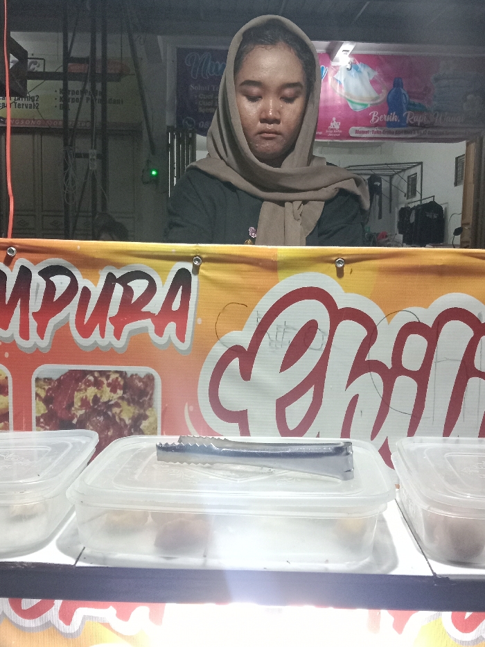 A medium-shot, front-facing image taken at night depicts a street food vendor in the city of Turunsih, Indonesia. The central figure is a woman, wearing a dark top and a light brown hijab, standing behind a brightly lit counter. Her eyes are partially closed or cast downwards, and her face exhibits a slight sheen. The counter is fronted by a colorful banner with red and white text, partially reading "MPURA" and "Ghili," alongside an image of food. On the counter, three clear rectangular plastic containers with lids are visible; one contains a pair of silver-colored tongs resting on its lid. The counter is illuminated from below by a strong artificial light source. In the background to the left, a dark building structure with visible wires and a partially visible sign with Indonesian text is present, along with a small green indicator light on a wall. To the right, another banner with purple and pink elements advertising "Nuansa" and "Bersih Rapi Wangi" (Clean, Tidy, Fragrant) hangs, behind which a metal laundry rack with clothes is discernible. The overall environment suggests an urban setting at night, likely a street or market area.