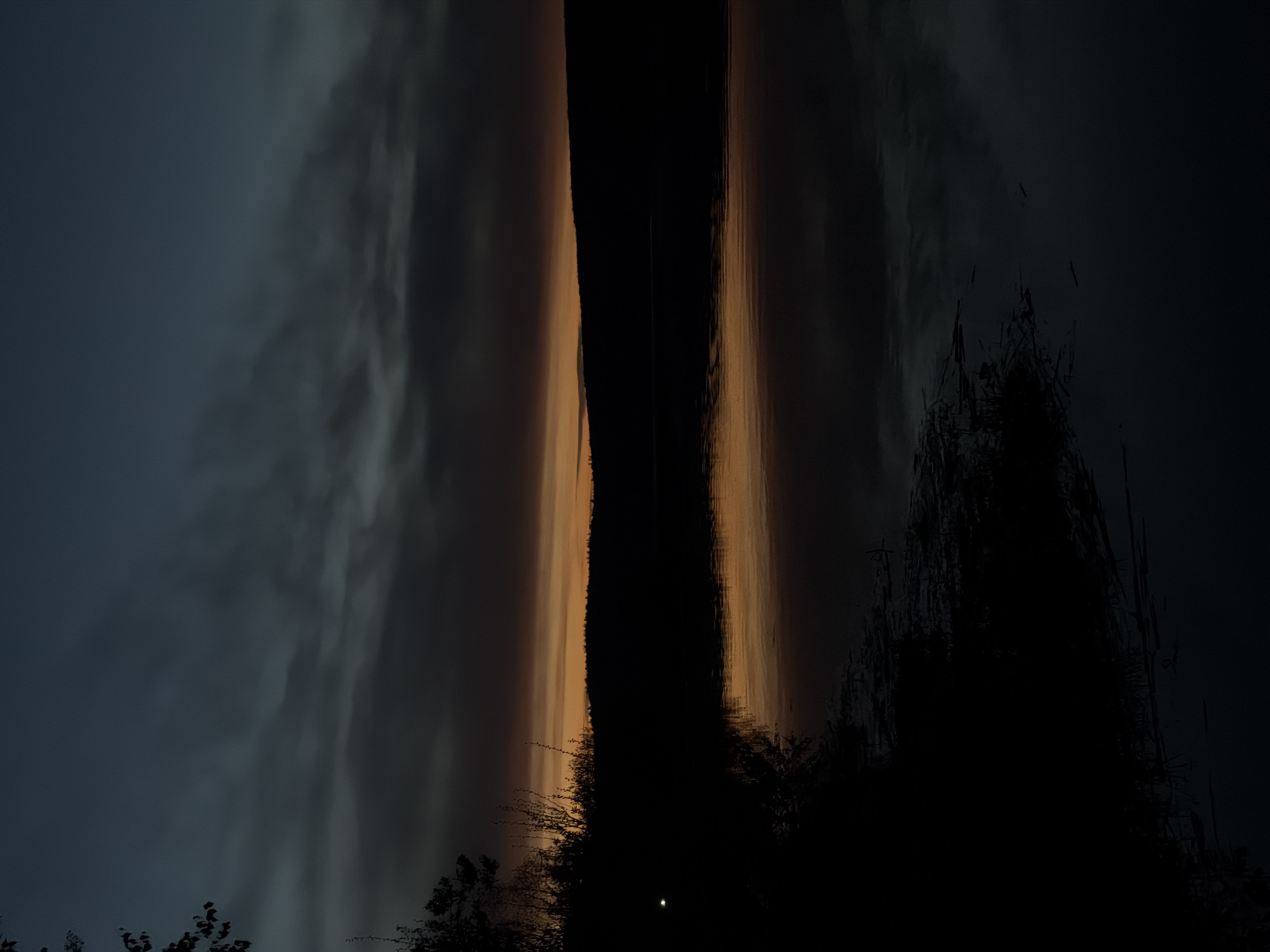 The image shows a dark, abstract view likely taken at night or in low light, possibly in Chaikovsky, Russia. Two light-colored columns are seen on either side of a very dark central space. The columns appear to be reflections on water, with the sky and trees reflected. The bottom of the image is obscured by dark silhouettes of trees or vegetation. A single light, potentially a distant light source or reflection, is visible near the bottom. The environment is dark and possibly wet, given the reflective surfaces.