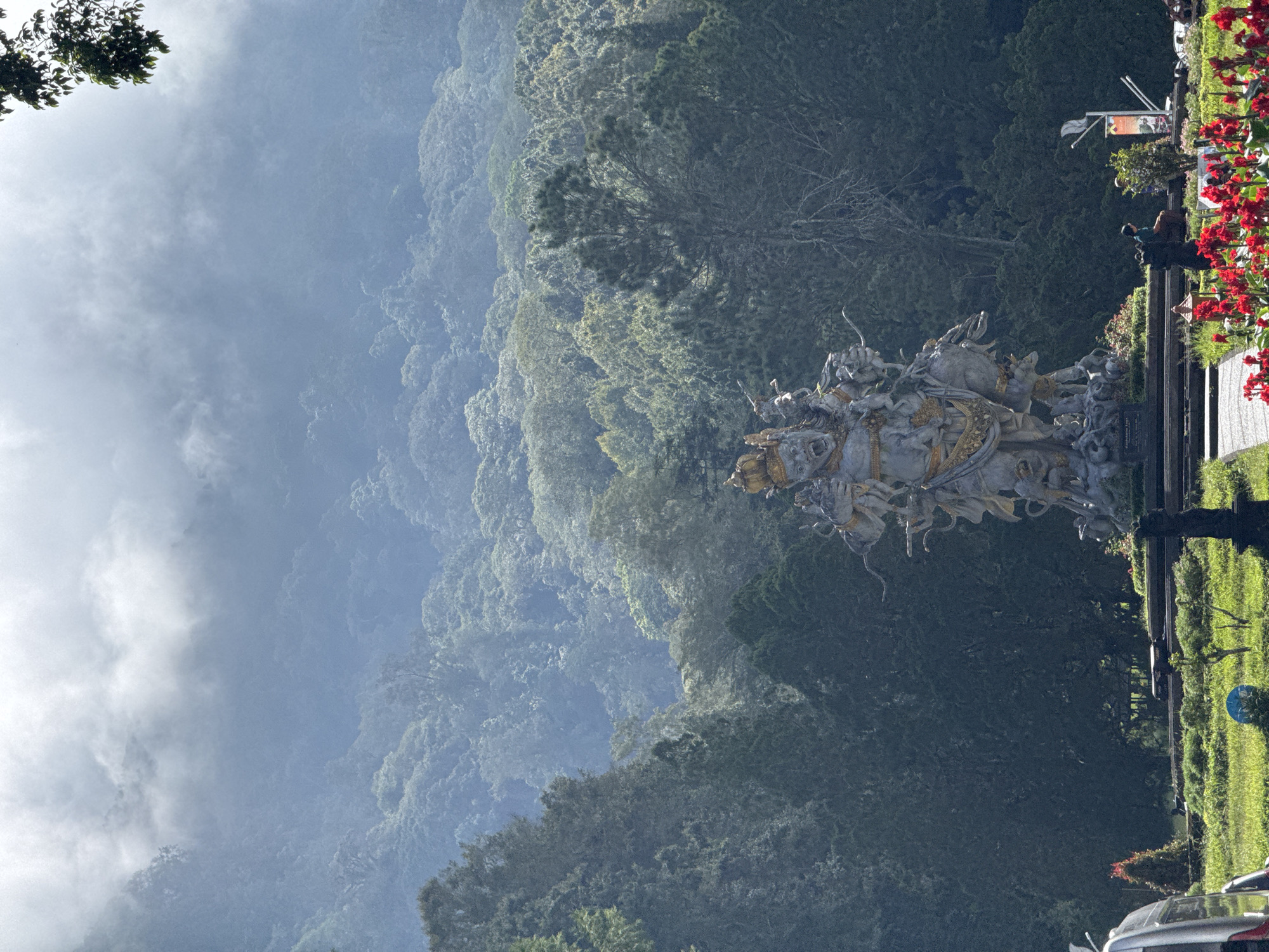 The image shows the Ghatotkaca statue in Baturiti, Bali, Indonesia. The statue is visible in the foreground, depicting a mythological scene with figures and horses. The background consists of a lush, green forest cascading down a mountain, partially obscured by mist or clouds. The sky is overcast, contributing to a hazy atmosphere. To the right of the statue, some greenery, red flowers, and parked cars are visible, indicating the statue's location within a developed area. A person is standing near the railing close to the flowers.