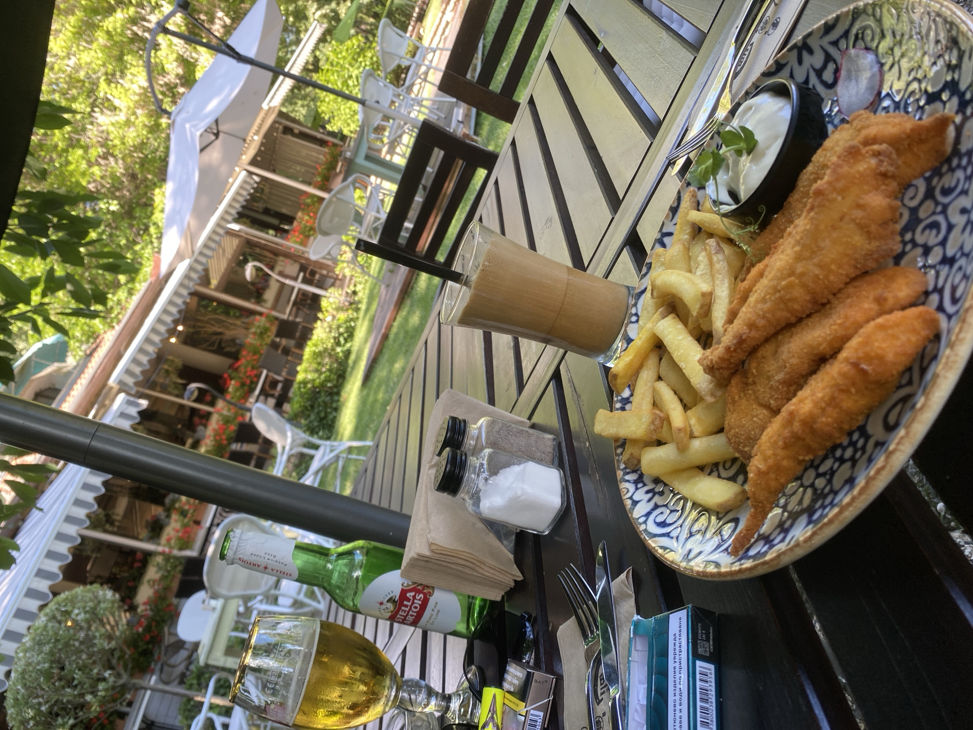 An outdoor seating area of a restaurant is depicted with a table set for a meal. On the table, there is a glass of beer, a bottle of Stella Artois, a glass of iced coffee with a straw, a salt and pepper shaker, a pack of cigarettes, cutlery, and a plate of food containing fried fish strips and french fries with a small bowl of dipping sauce. The table is made of wooden planks, and in the background, there are more tables and chairs, umbrellas, lush greenery, and the facade of a building with awnings. The setting appears to be a sunny day.