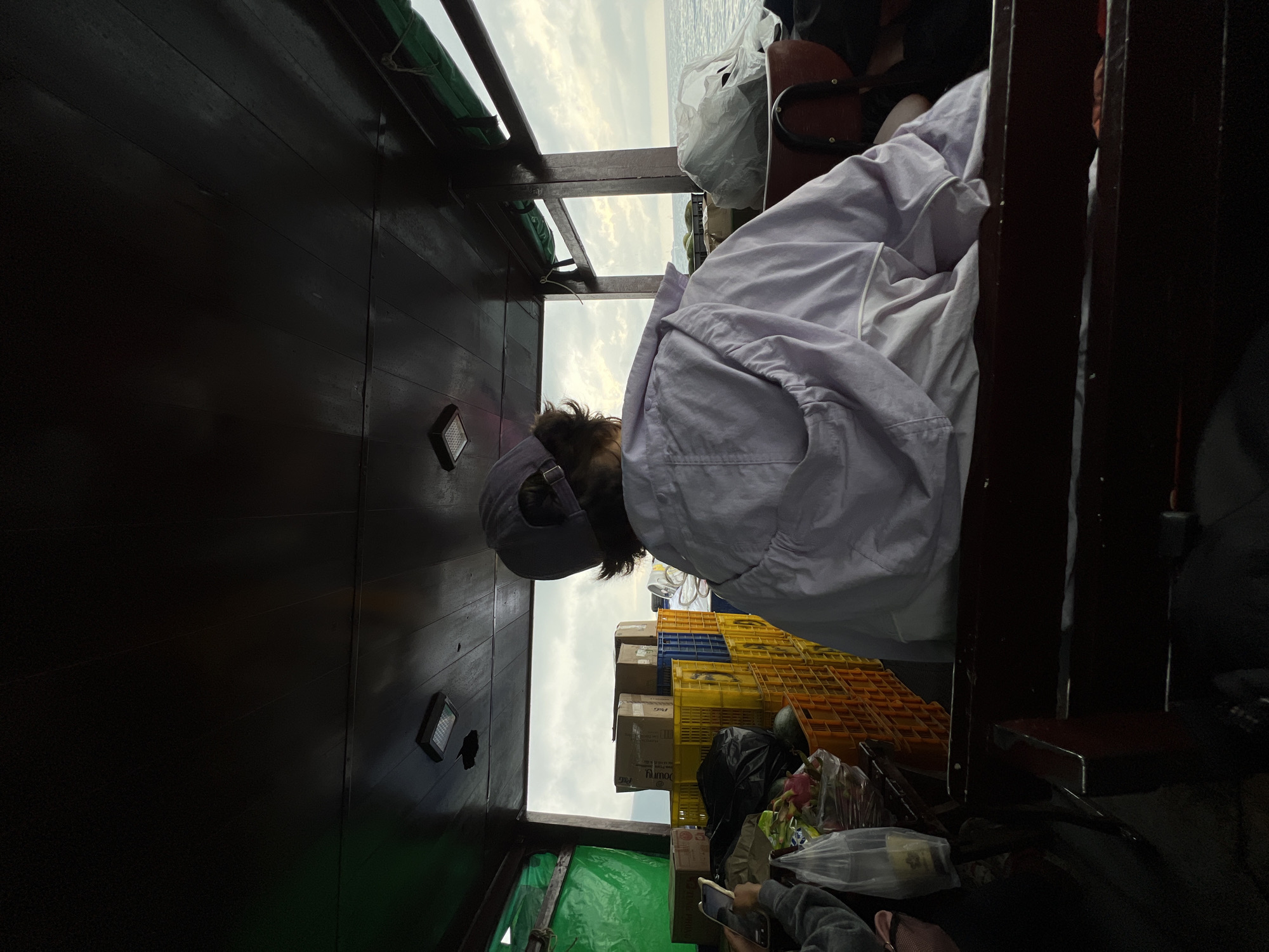 A person wearing a grey baseball cap and a white jacket is on a boat, possibly a fishing vessel, judging by the crates of fish stacked up. The boat has dark wooden planks on the side, and the view outside shows water and a cloudy sky. The location is likely Bac Nha Trang Ward, Vietnam, based on the description. The person's face is not visible, and they appear to be either seated or standing. There are also various bags and objects piled up in the boat. Someone is also taking a picture with a cellphone in the left corner of the frame.
