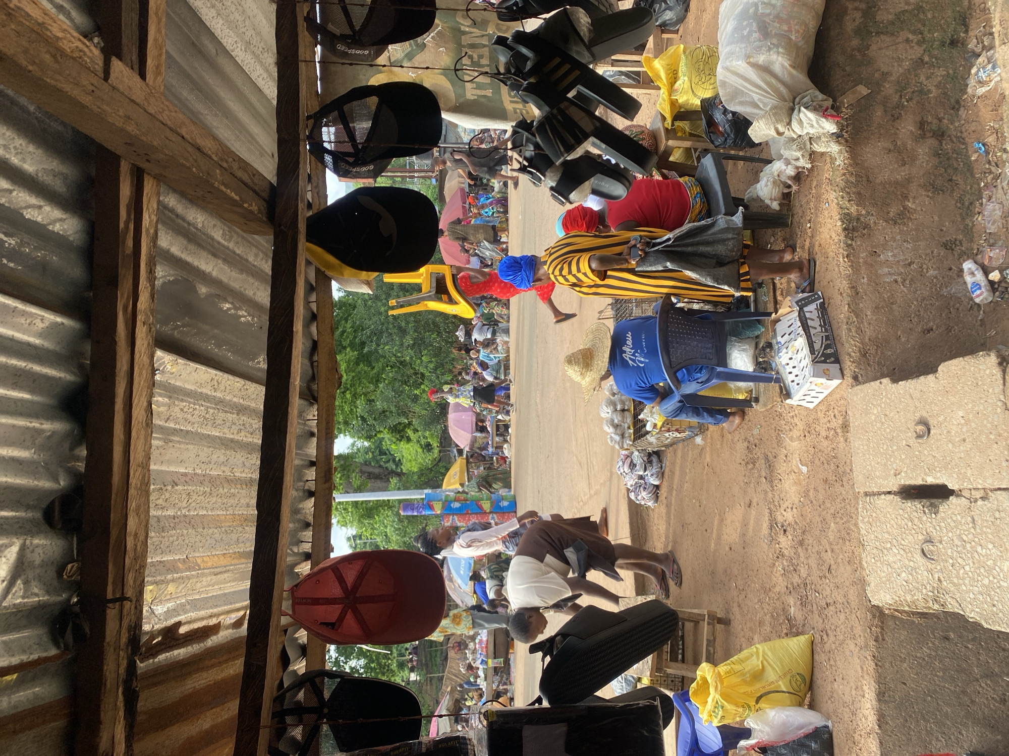 The image depicts a bustling street scene in Igbara, Nigeria, viewed from within a corrugated iron structure, possibly a shop or stall.
People are seen walking along the street, with umbrellas providing shade. Various objects are displayed, including hats, chairs, and bags. Some individuals are seated, possibly engaged in buying or selling activities. Debris is visible on the ground near the corrugated iron structure. The overall environment suggests an active marketplace or commercial area.