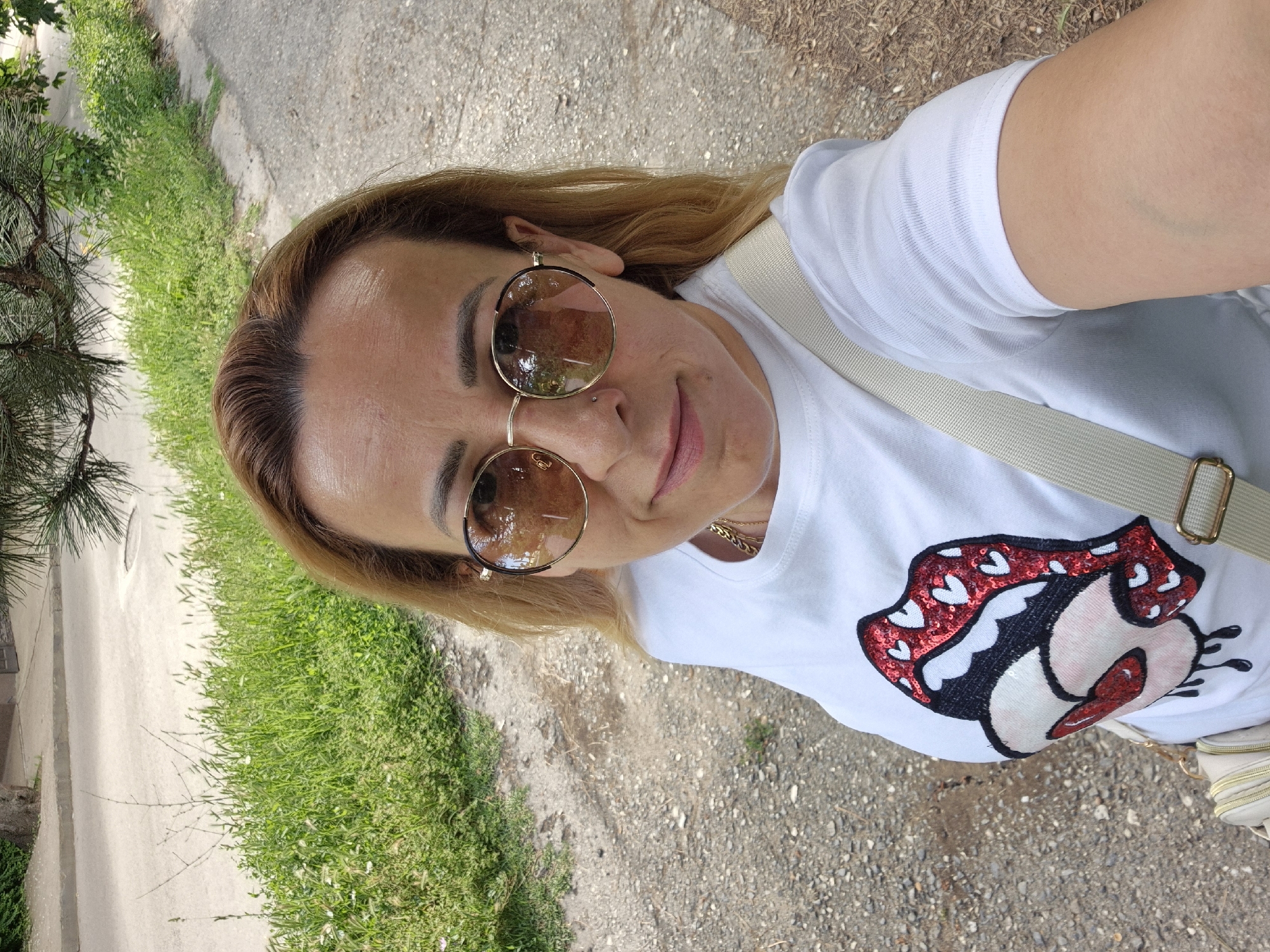 An outdoor, medium shot selfie of a woman with blonde hair and sunglasses. She is wearing a white t-shirt with a sequined red and white lip graphic. A light-colored strap of a purse is visible across her chest. The background shows a paved pathway bordered by green grass and trees.