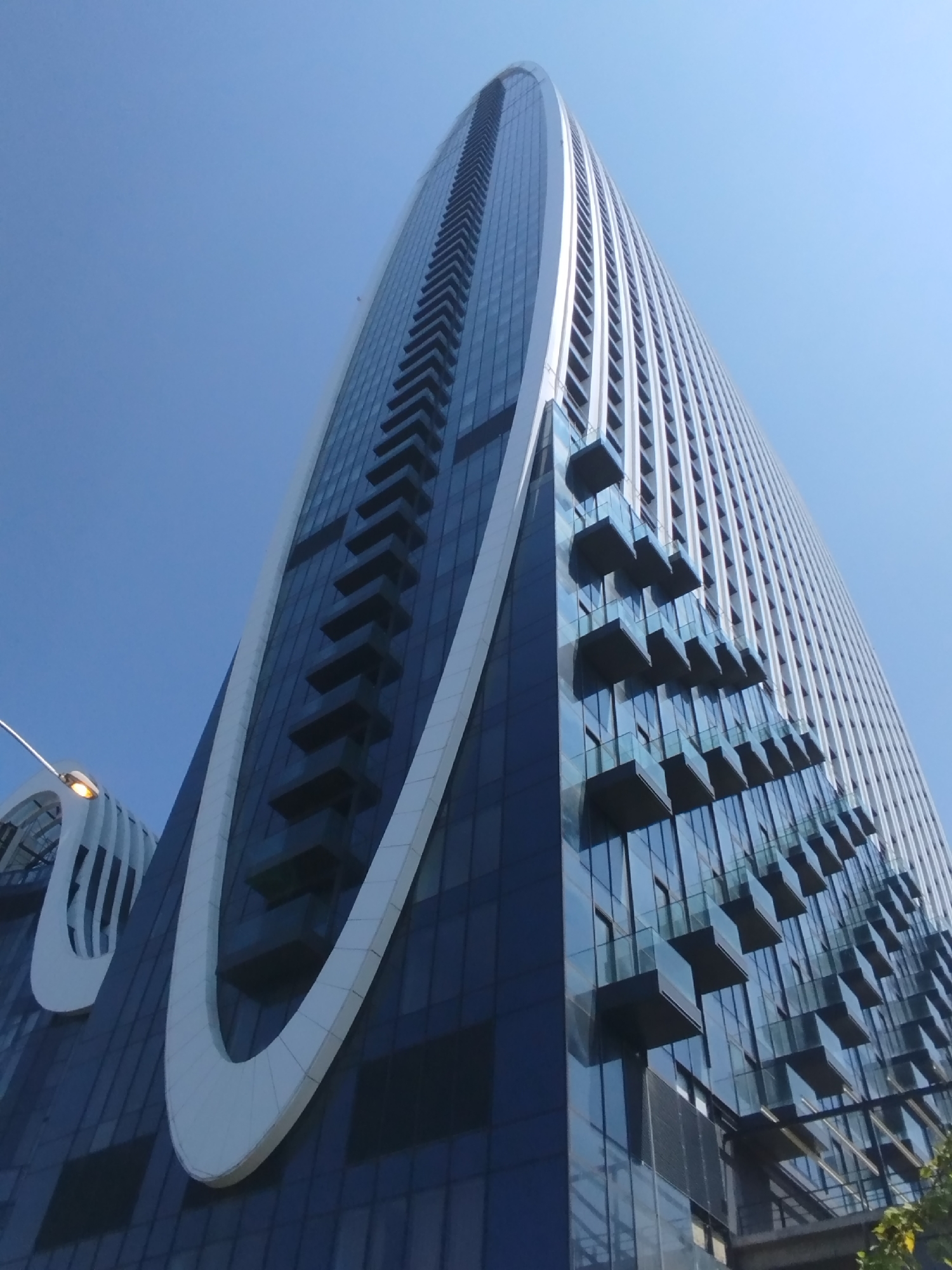 The image is an upward-angle, full shot of a modern skyscraper in Batumi, Georgia, under a clear blue sky. The skyscraper has a distinctive, tall, oval-shaped white structure that wraps around a portion of its facade. The building is predominantly clad in dark blue glass, with rows of balconies extending outwards at various levels, each featuring glass railings. The balconies appear to be protruding from recessed sections of the building's exterior, creating a stepped effect. On the left side of the image, a curved, white architectural element is visible, also with a glass-fronted section below it, and a street lamp with a warm light is illuminated. The perspective emphasizes the height and sleek, contemporary design of the building.
