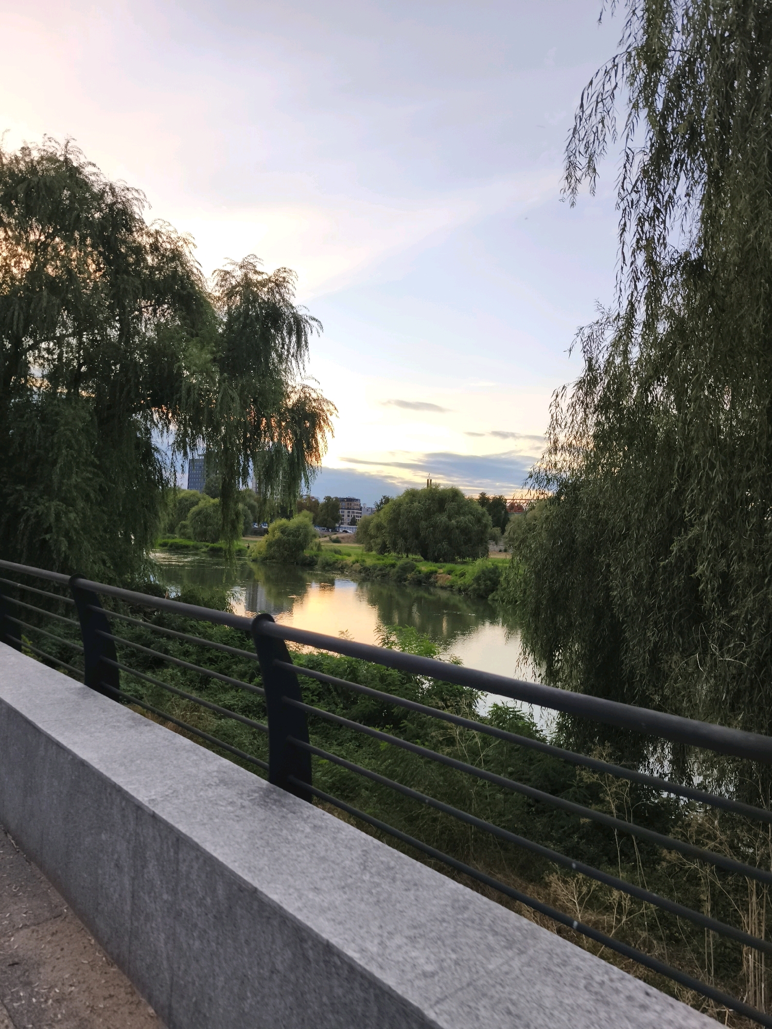A landscape image shows a river with trees and buildings on the opposite bank. The sky is partly cloudy and the sun is setting. A stone wall and metal railing are in the foreground. The scene appears to be in a park or along a riverwalk.