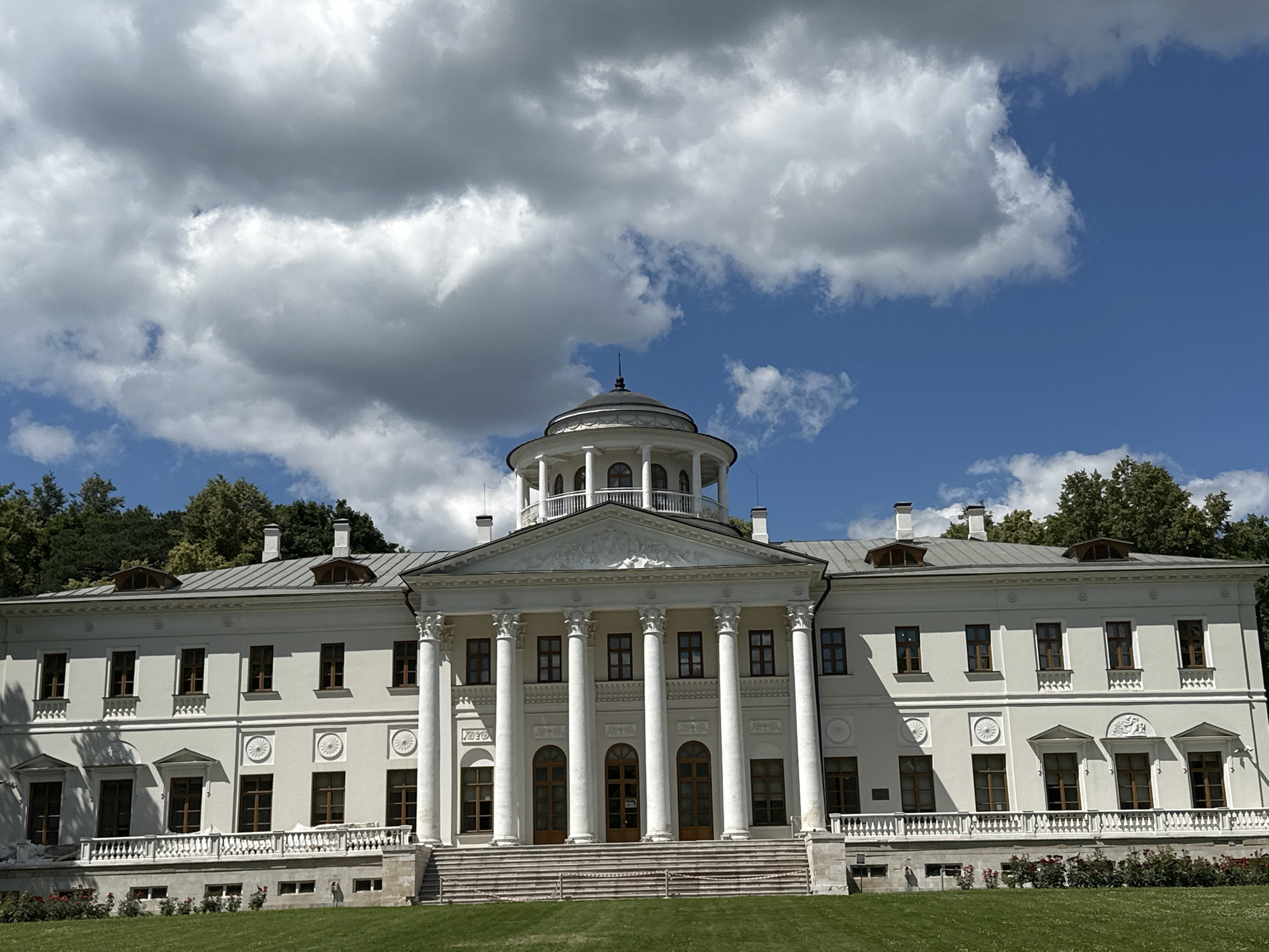 The image shows a grand, white neoclassical building with a prominent portico supported by tall columns. The building has a symmetrical facade with many windows and ornate architectural details. A circular, multi-story structure topped with a dome is visible in the center, above the portico. The building is set against a backdrop of a bright blue sky filled with dramatic, white and grey clouds. Lush green grass covers the foreground, with a row of red flowers visible along the base of the building. Trees are present on a slight incline behind the structure. The scene appears to be an exterior shot of the State Museum "Ostafyevo", the Russian Parnassus, located in Ostafyevo, Russia. There are no people or animals visible in the image, and no actions are taking place. The overall impression is one of stately architecture and a pleasant outdoor environment.