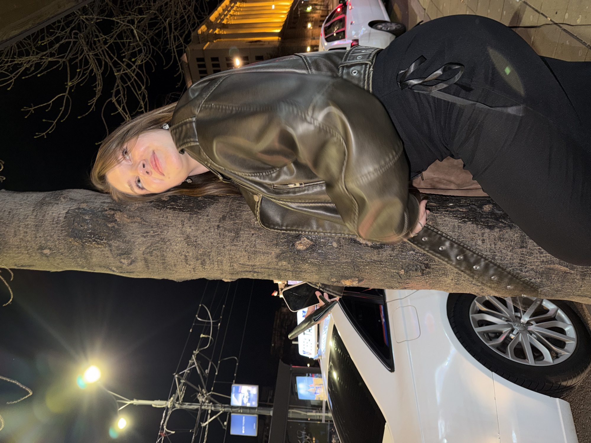 A young woman is leaning against a tree at night in Belgrade, Serbia. She is wearing a leather jacket and black pants with a bow at the waist. A white car is parked nearby, and there are city lights and buildings in the background. The sky is dark, and bare tree branches are visible.