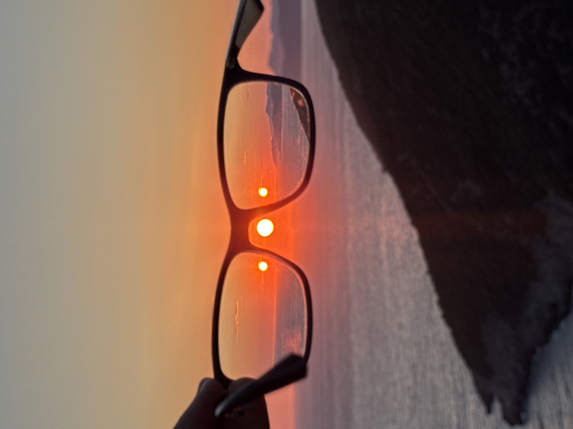 A hand holds a pair of eyeglasses in front of a sunset over the Adriatic Sea. The sun, a vibrant orange orb, is visible through both lenses, appearing as two distinct circular reflections. The sky transitions from a soft orange at the horizon to a pale blue at the top. The sea below is a deep purple, reflecting the sunset's colors. Silhouetted mountains rise in the distance, their peaks softened by the haze of the setting sun. In the foreground, the dark, rocky outline of a hill slopes down towards the water. The image was captured in the vicinity of Zagora, Montenegro.