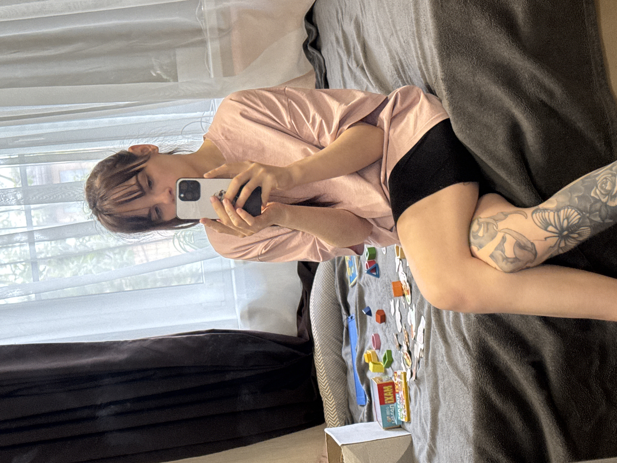 A woman lies on a bed next to a window with white curtains. She is wearing a pink shirt and black shorts and is holding a white cell phone up to take a picture. The woman has tattoos on her right arm. On the bed, next to the woman, are a box and several wooden and plastic toys. The location in the city of Rostov-on-Don, Russia cannot be determined from this photo.