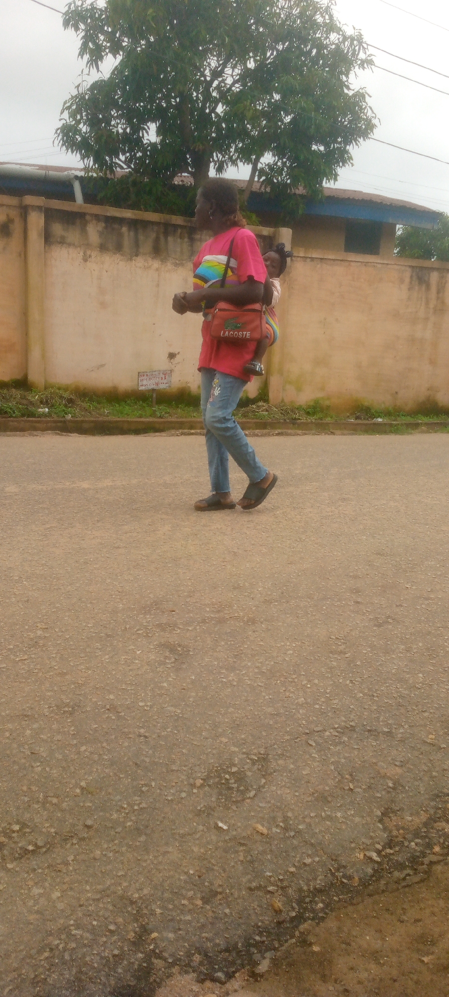 An adult with dark skin and dark hair is walking on a paved road in an urban environment. The adult is wearing a bright pink t-shirt with a multi-colored striped design across the chest and light blue jeans with an embroidered design on the right leg. A young child, also with dark skin and dark hair, is being carried on the adult's back, secured by a fabric wrap. The child is wearing a light-colored garment. A small orange bag with the "LACOSTE" logo is slung across the adult's front. In the background, there is a light-colored, weathered wall with a green grassy strip at its base. Beyond the wall, there are trees with green foliage and a building with a dark roof. Overhead, power lines are visible against a cloudy sky. The road surface is composed of rough, light brown asphalt. The scene is captured from a side-view perspective, with the adult and child in the mid-ground.