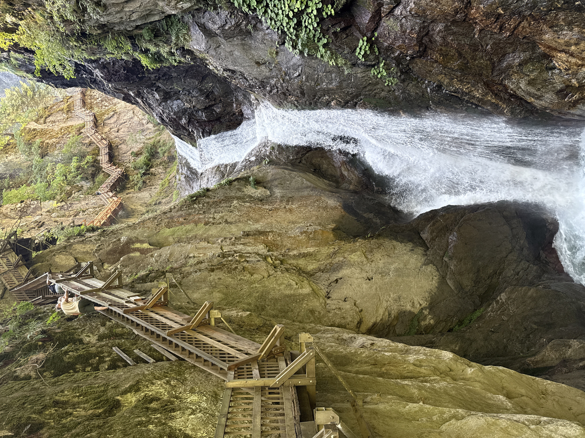 The image depicts the Gorge du Trient, a spectacular natural canyon located in the municipality of Trient, within the canton of Valais, Switzerland, near Martigny-Combe. The scene is dominated by a cascading waterfall plunging into a rocky gorge. Wooden walkways and staircases are constructed along the steep canyon walls, allowing visitors to navigate the dramatic terrain and experience the waterfall up close. In the foreground, two individuals are ascending one of the wooden staircases, their figures dwarfed by the sheer scale of the natural environment and the man-made structures. Vegetation, including moss and small plants, clings to the rocky surfaces. The sunlight filters through the gorge, illuminating parts of the rock faces and the mist rising from the waterfall.