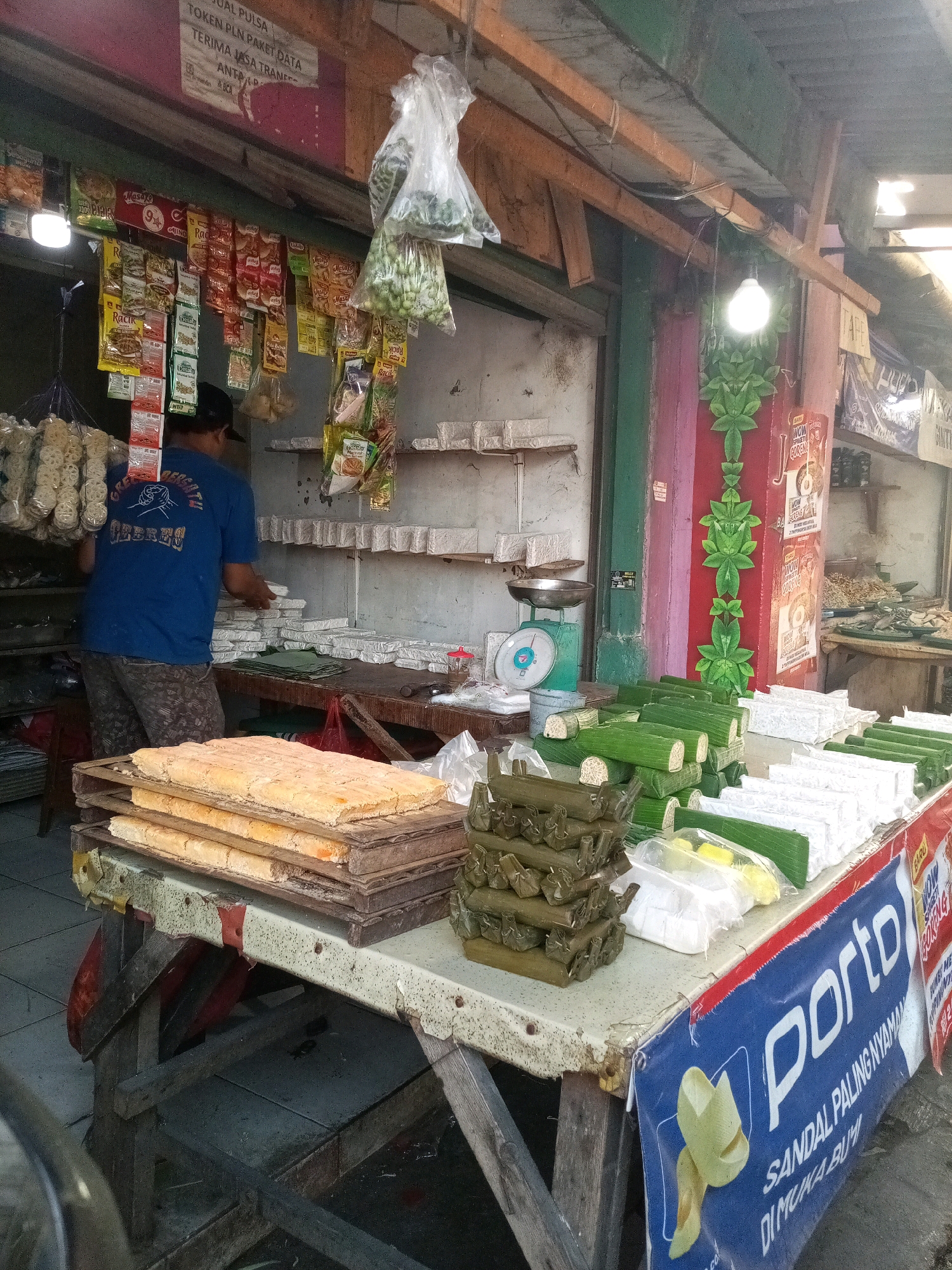This image depicts a street-side food stall in Bekasi, Indonesia, likely a traditional market. In the foreground, a table is laden with food items, predominantly tempeh wrapped in banana leaves. These are arranged in stacks of cylindrical bundles and rectangular blocks. To the left, on a wooden pallet, lie several large, square blocks of what appears to be fried tofu or tempeh, stacked on top of each other. A person, seen from behind, wearing a blue t-shirt and patterned shorts, is attending to the stall, possibly arranging the food. Behind the table, shelves are stocked with more food products and packaging materials. Bags of snacks and spices hang from the ceiling. A weighing scale sits on a shelf. The stall's structure includes a wooden roof and colorful painted pillars. A blue banner with white text and an illustration of yellow fruit is attached to the front of the table. The overall environment suggests a bustling, open-air market setting.