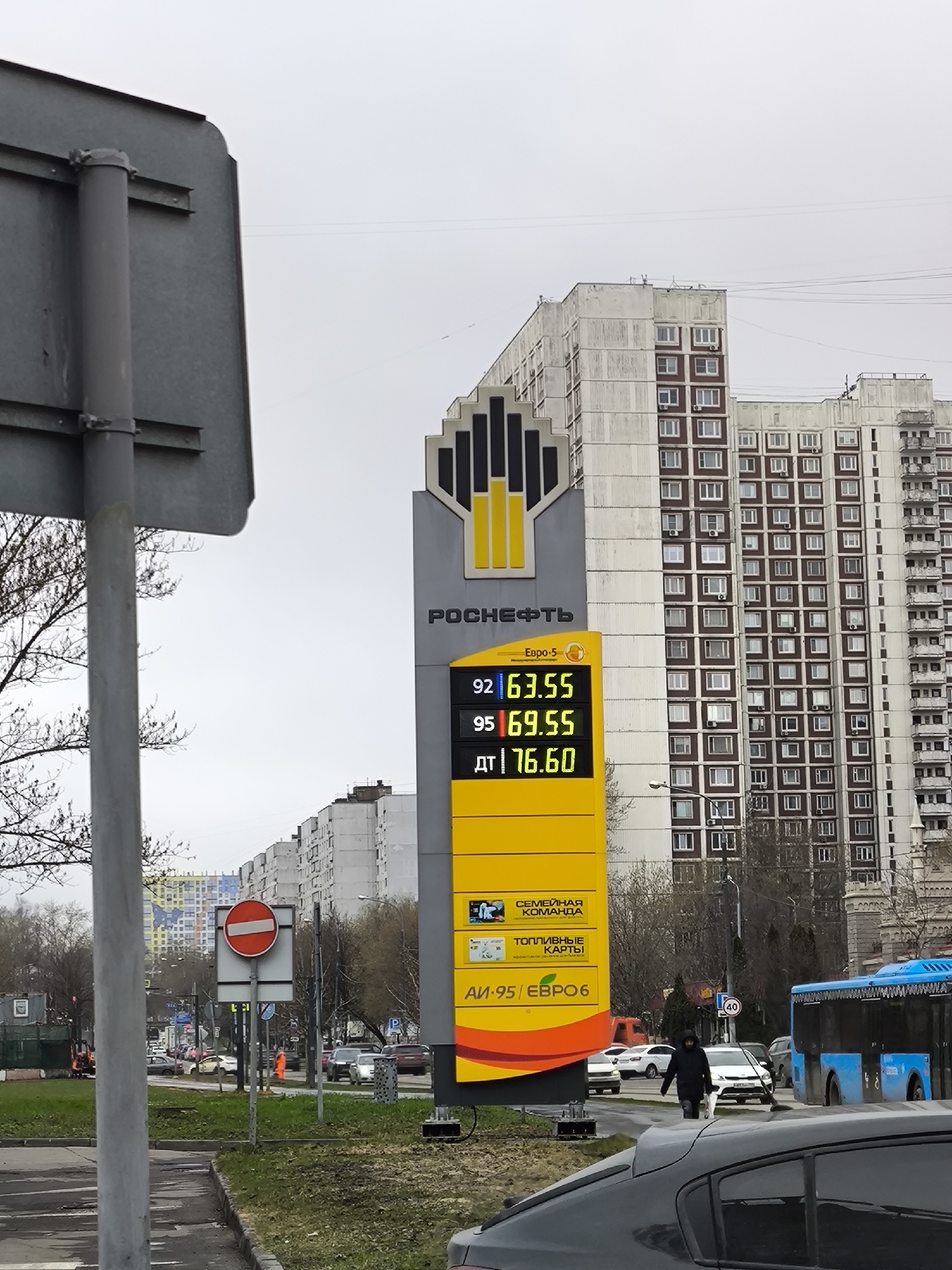 The image shows a Rosneft gas station in Moscow, Russia. The sign displays the current prices for different grades of fuel: 92 octane at 63.55, 95 octane at 69.55, and diesel at 76.60. The building behind the sign is a large, multi-story apartment building. In the foreground, there is a street with cars and a blue bus, and a person crossing the street. There is a 'no entry' traffic sign on a grass median. The sky is overcast.