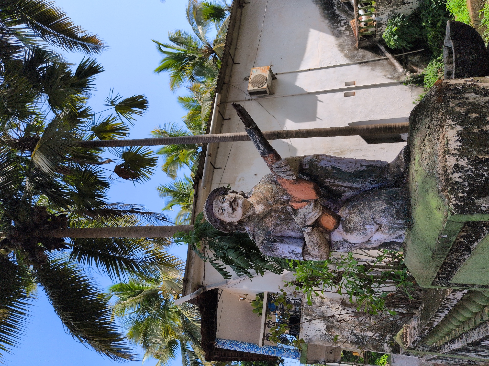 A weathered statue of a man in a hat and coat, playing a stringed instrument, kneels on a moss-covered pedestal. The statue is the primary subject, located in the foreground, and is a focal point of the image. Behind the statue, a white building with a corrugated metal roof is visible, featuring an air conditioning unit mounted on the exterior wall. Palm trees, with lush green fronds, dominate the background, indicating a tropical or subtropical environment. The scene is outdoors, under a clear blue sky. The overall impression is one of a weathered, possibly aged, outdoor setting with a rustic statue as a central element. The location is identified as Oxel, India.