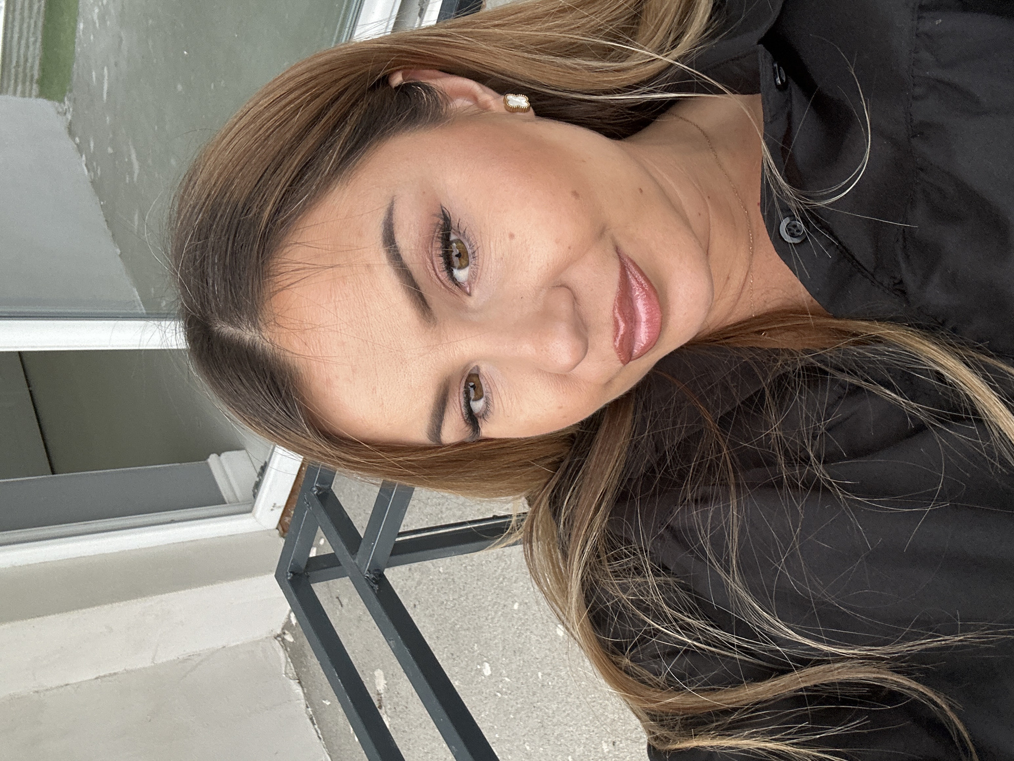 A medium shot selfie of a woman with long, brown and blonde hair, wearing a black collared shirt. She has on makeup, including winged eyeliner and lip gloss. She is wearing earrings. The woman is outdoors, with a building and railing visible in the background. It appears to be a cloudy day.