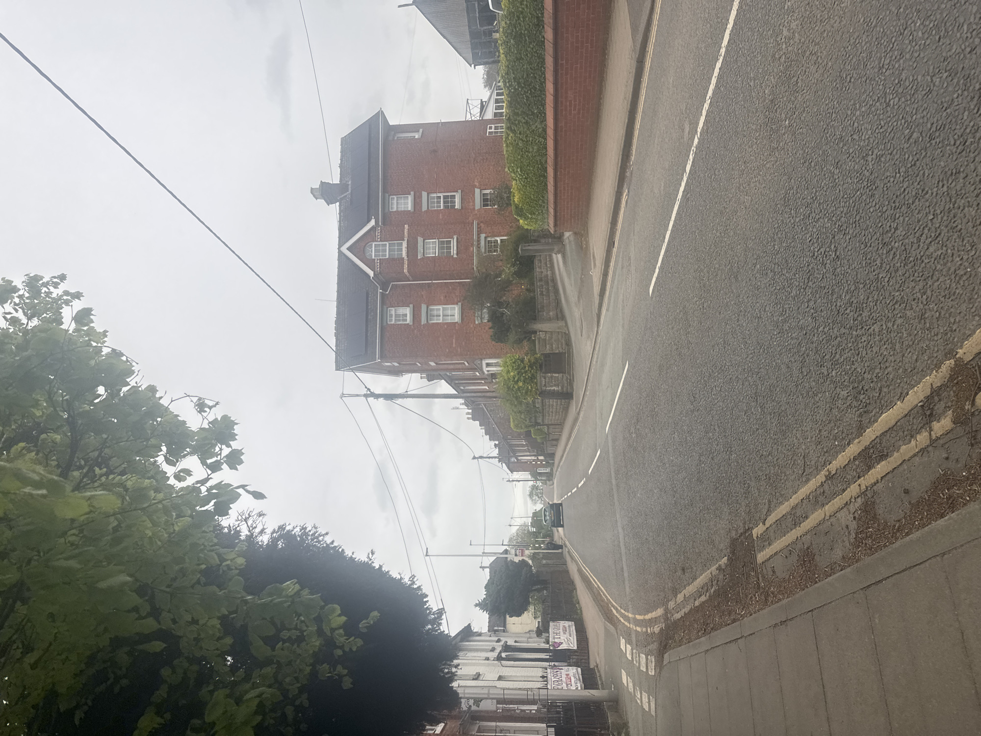 This image depicts a quiet street scene in Welshpool, United Kingdom, under an overcast sky. The photograph is taken from a tilted perspective, showing the street ascending diagonally from bottom right to top left.
On the right side, an asphalt road features clear white dashed lines and double yellow no-parking lines along the curb. A grey pavement runs parallel to the road, bordered by a low brick wall adorned with lush green ivy or shrubbery. Behind this, a substantial red brick building stands prominently, characterized by multiple white-framed windows, including an arched window on a gable, and a dark, possibly tiled roof with some dark panels (potentially solar panels).
The upper part of the frame is dominated by a pale, cloudy sky crisscrossed by several overhead utility lines and poles. In the lower left, dense green foliage from trees partially obscures the view of the street and buildings beyond. Further down the street on the left, a row of residential buildings, some painted white, are visible. A dark-colored car is parked further in the distance on the left side of the road.
Visible text on signs along the left pavement includes "THE ORCHID" and directly below it, "PARAYAN'S." The overall weather appears dull and grey, indicative of an overcast daytime. There are no people visible, and no specific activity is taking place, presenting a serene, almost deserted urban landscape.