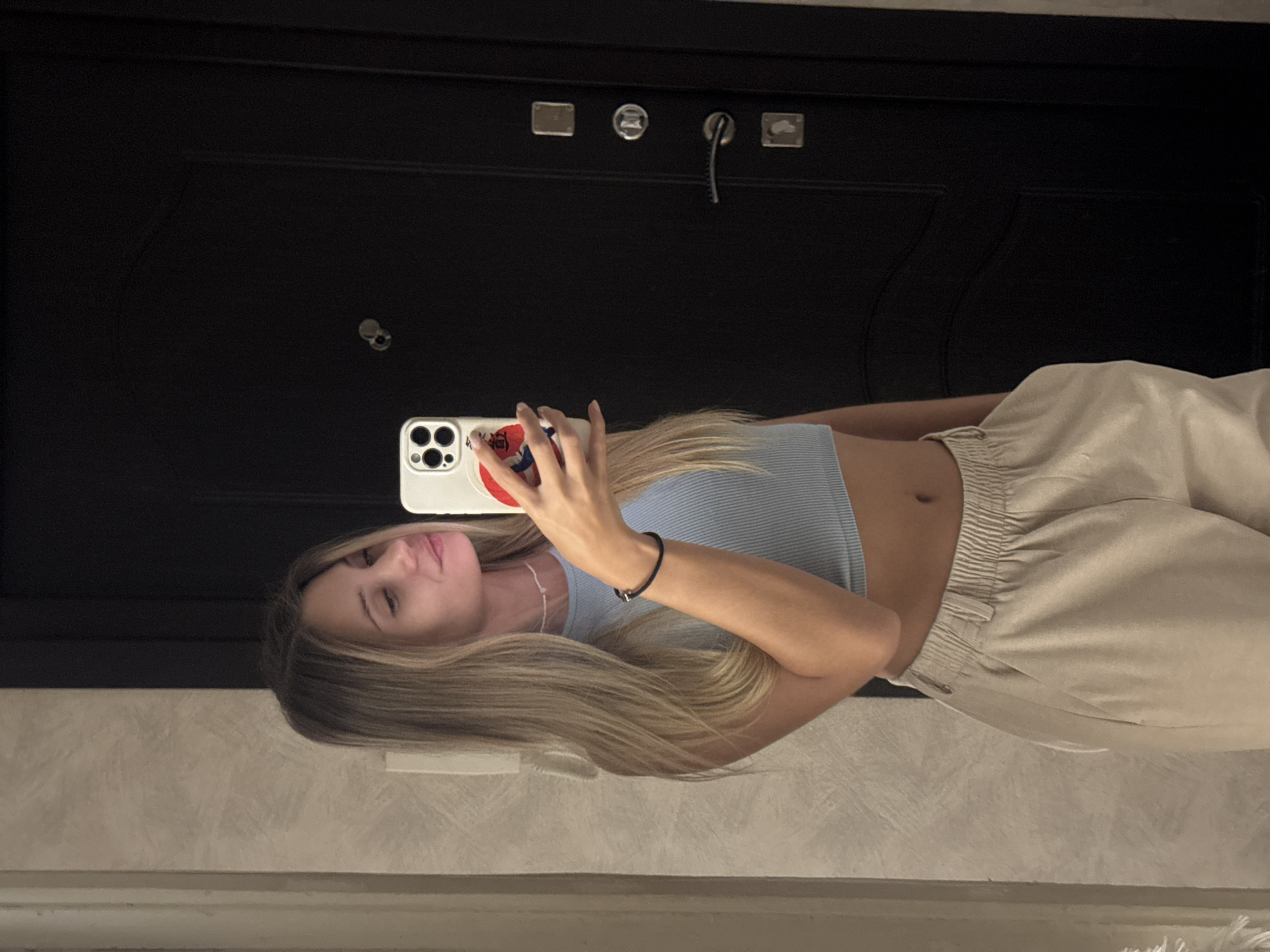 A young woman is depicted taking a mirror selfie indoors. The image, originally rotated 90 degrees counter-clockwise, shows her facing forward, holding a white smartphone with multiple camera lenses in her right hand, capturing her reflection.
She has long, straight blonde or light brown hair draped over her shoulders. She is dressed in a light blue ribbed crop top, exposing a small portion of her midriff, and light-colored (beige or tan) sweatpants with an elasticated waist. A black bracelet is visible on her left wrist, and she wears a thin white necklace. Her expression is neutral to slightly smiling.
The background features a dark, possibly black or dark brown, door. On the door, several metallic fixtures are visible: two rectangular plates, a round lock mechanism, and another lock or handle from which a black cord or wire descends. The lower part of the image shows a light-colored, possibly textured, wall or floor. No discernible text is present in the image. The setting appears to be an indoor environment, likely a hallway or room, under artificial lighting.