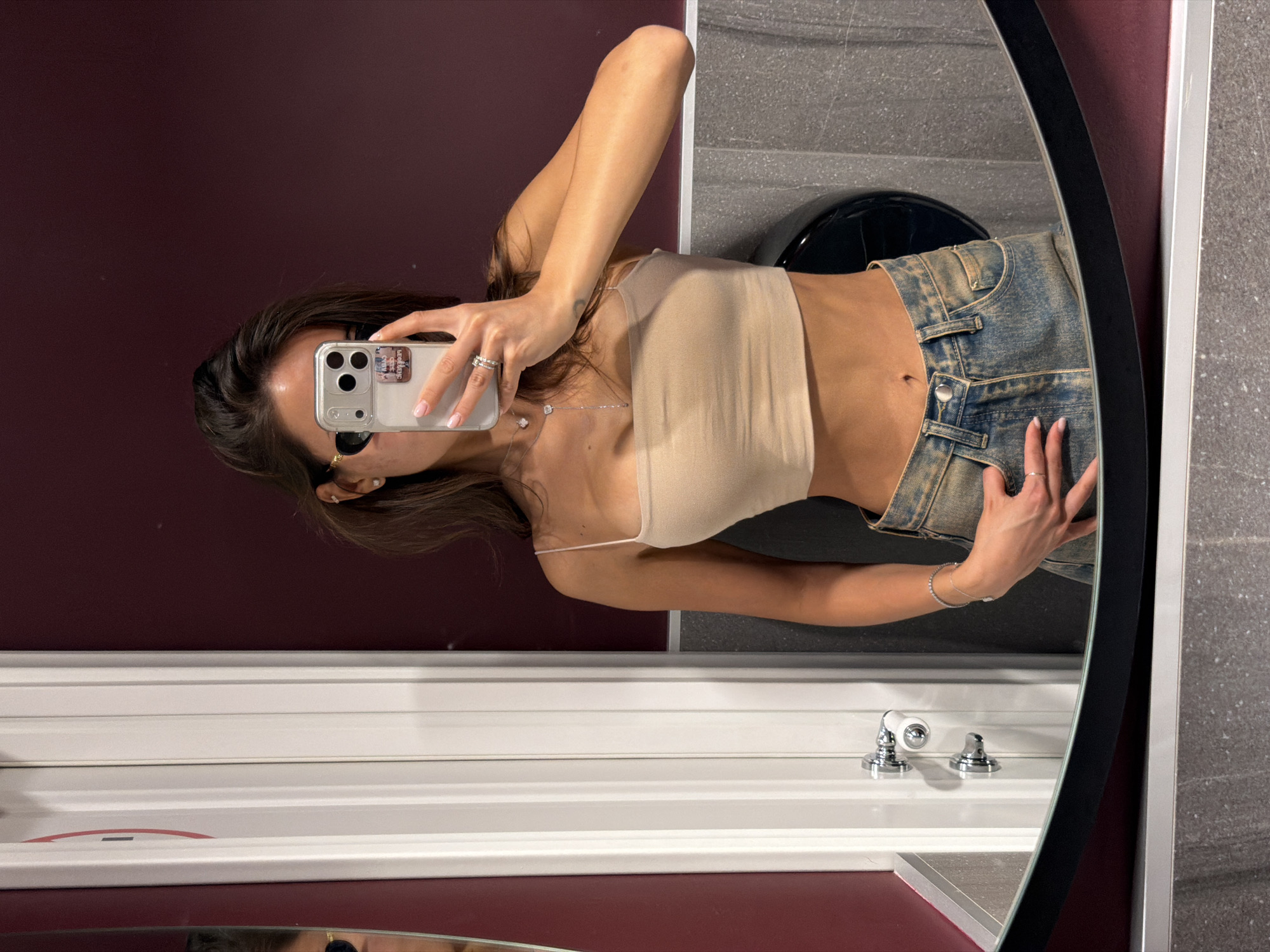 A woman in a tan top and blue jeans is taking a selfie in a bathroom mirror. The bathroom has maroon walls, a white countertop, and a gray stone wall. A black toilet is visible in the background. The location is likely in Krasnoyarsk, Russia.