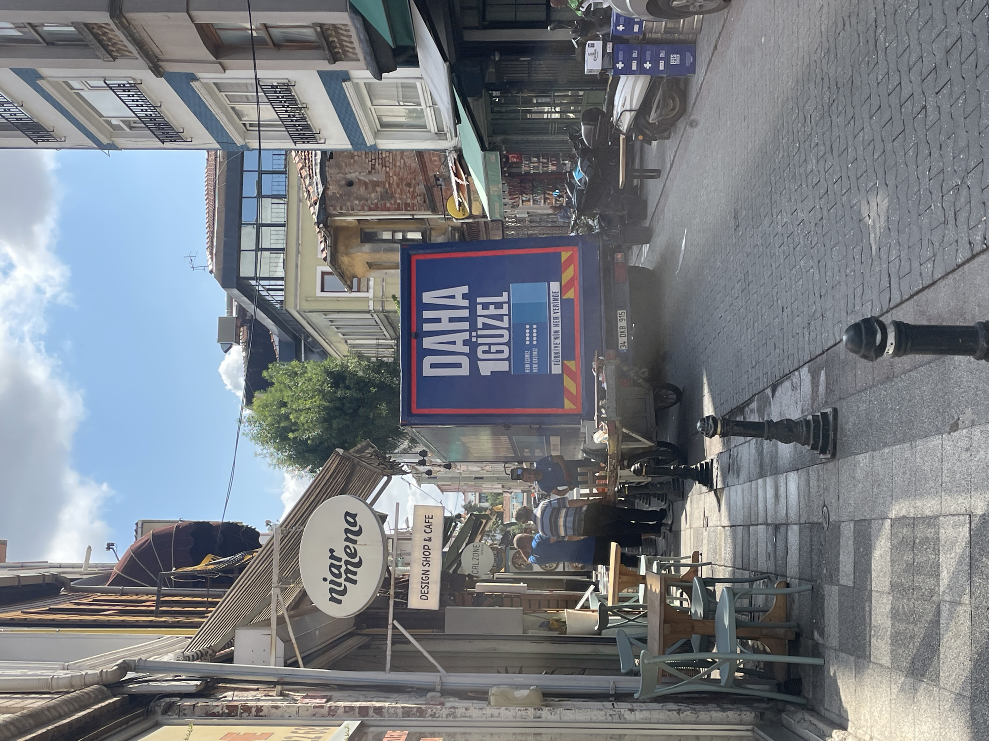 The image captures a sunlit street scene in Kadıköy, Turkey, with buildings lining both sides of a cobblestone road. A blue truck with large white text that reads "DAHA 1 GÜZEL" is parked in the middle of the street. To the left, an outdoor seating area for a cafe is visible, with tables and chairs. Several individuals are present, some standing near the truck and others near the cafe entrance. The environment suggests a bustling urban setting with commercial establishments and pedestrian traffic.