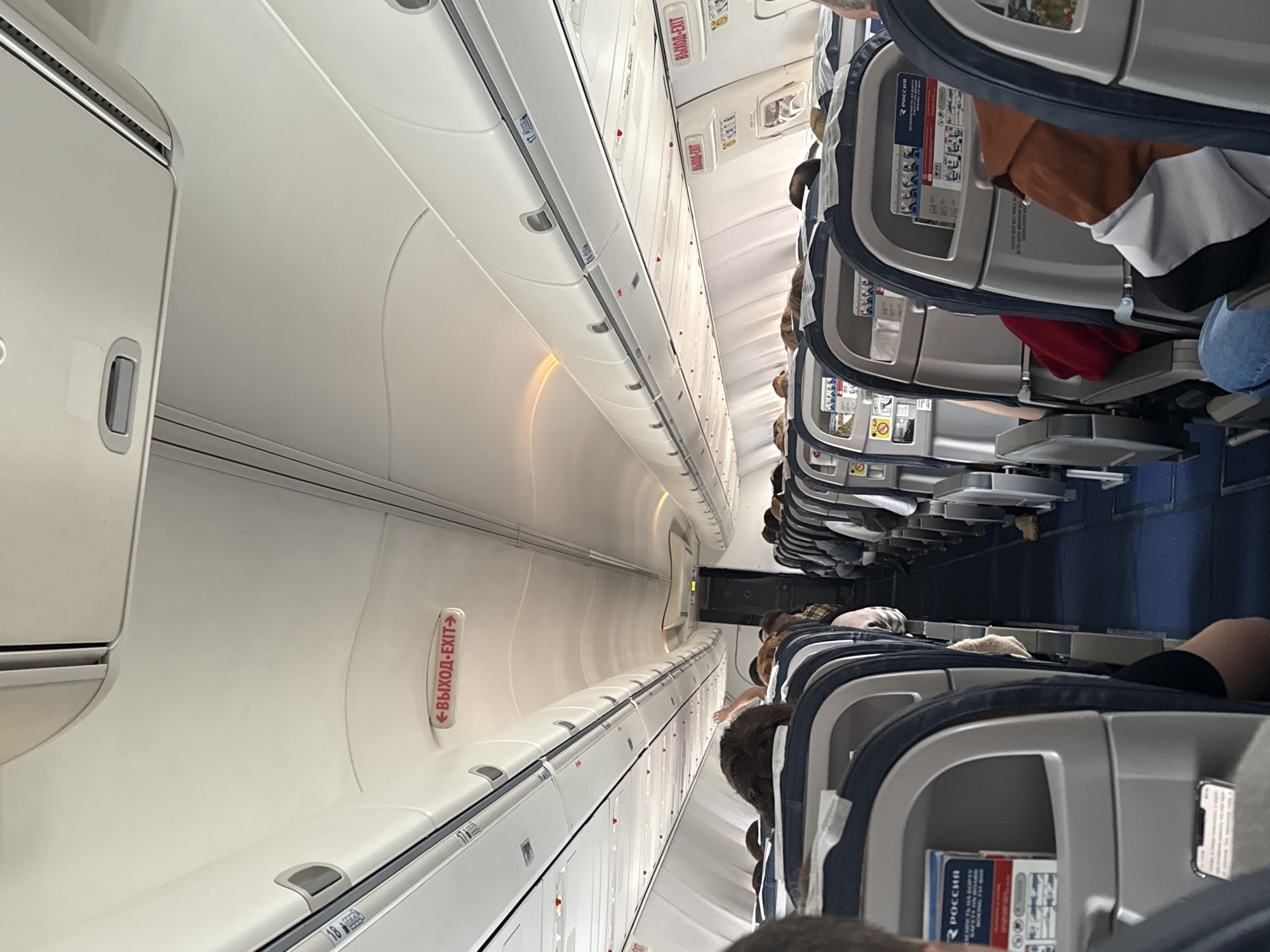 The scene depicts the interior of a commercial passenger aircraft cabin, identified as a Boeing 737-800 operated by Rossiya Airlines, presumably associated with operations at Ufa Airport, Russia. The view is from a seated passenger's perspective, looking towards the rear of the aircraft down a central blue-carpeted aisle. Overhead white paneling and closed luggage compartments line both sides of the cabin. Multiple illuminated red "ВЫХОД-EXIT" signs are visible along the overhead panels, some with directional arrows. Numerous passengers are seated in the grey and blue upholstered seats, predominantly facing forward. In the immediate foreground, a smartphone is partially visible in a seatback pocket on the left. Further down the left aisle, a passenger is seen with an arm raised. Safety information cards bearing the "РОССИЯ" logo are present in the seatback pockets of several seats. Artificial interior lighting illuminates the cabin.