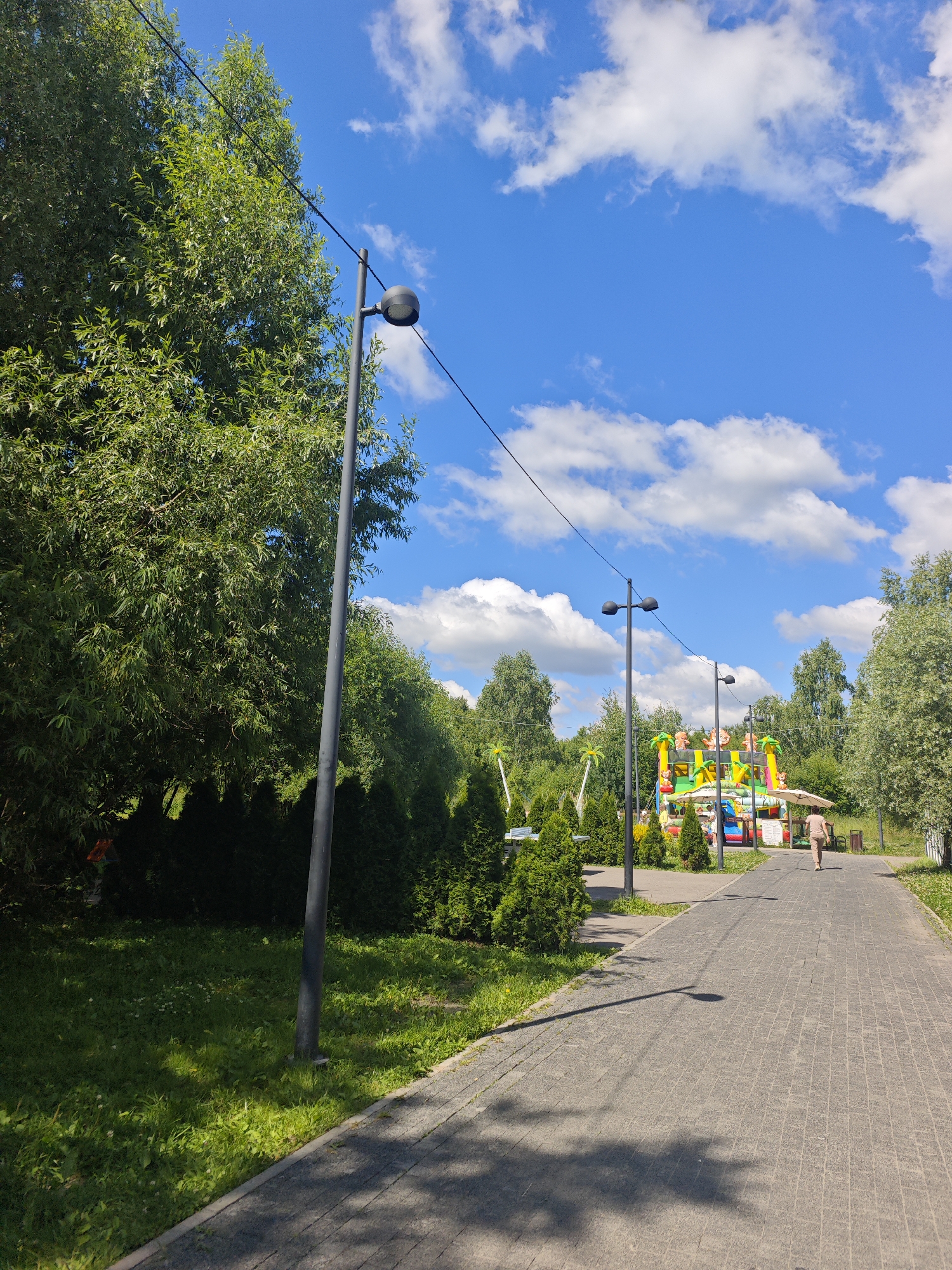 The image depicts a paved walkway in a park on a sunny day. Tall green trees line the left side of the walkway, with a grassy area in front of them. Several lampposts are visible along the path. In the distance, a colorful inflatable playground structure with cartoon animals is set up, suggesting it's a recreational area. A lone person, a woman wearing a light brown outfit, is walking away from the camera on the walkway, heading towards the playground. The sky is bright blue with scattered white clouds. The scene appears to be in a park or amusement area within the city of Chekhov, Russia, given the context of the location provided.