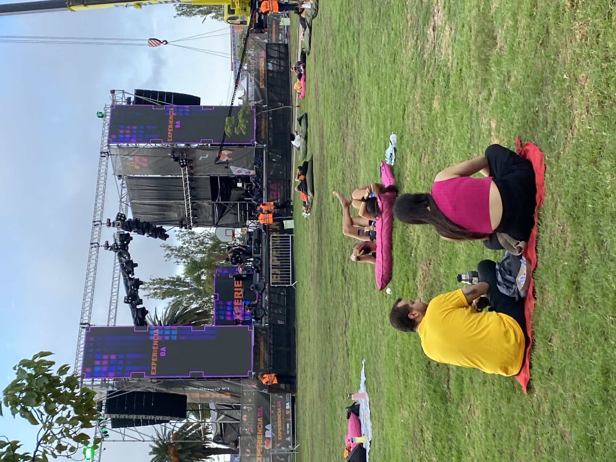 The image depicts a daytime outdoor event, possibly a concert or festival, taking place on a grassy area in Buenos Aires, Argentina. A large stage structure is visible in the background, equipped with large screens displaying the words "EXPERIENCIA BA". A yellow crane is visible above the stage. People are scattered across the grass, some sitting on blankets and enjoying the event. Specifically, in the foreground, a man wearing a yellow shirt and black pants is seated on a red blanket. A woman wearing a pink top and black pants is sitting beside him on the same blanket.
