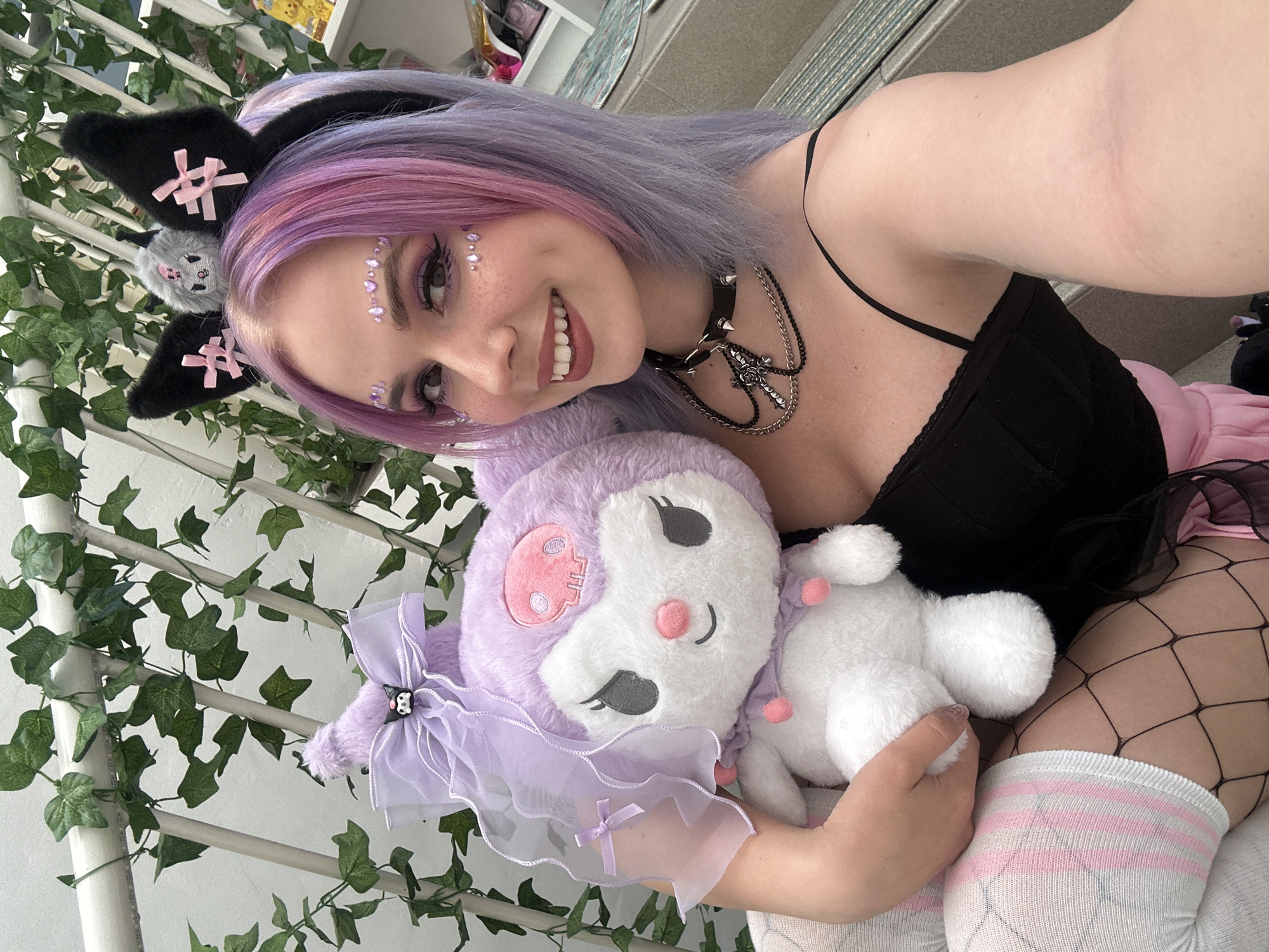 A young woman with lavender and pink hair, wearing cat ear headbands and facial jewel embellishments, is holding a stuffed Kuromi toy. She is wearing a black top, a spiked collar with a cross pendant, and fishnet stockings with striped thigh-high socks. The background includes greenery and white railing suggesting an indoor setting with plants. There is no information in the image to suggest it was taken in City of Zagreb, Croatia.